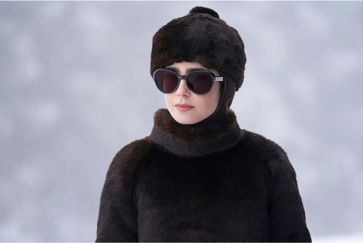 Pessoa vestindo um casaco e gorro de pele sintética em tons escuros, óculos de sol grandes estilo retrô e fundo nevado. Tendência de moda inverno, estilo sofisticado e aconchegante, ideal para proteção contra o frio.