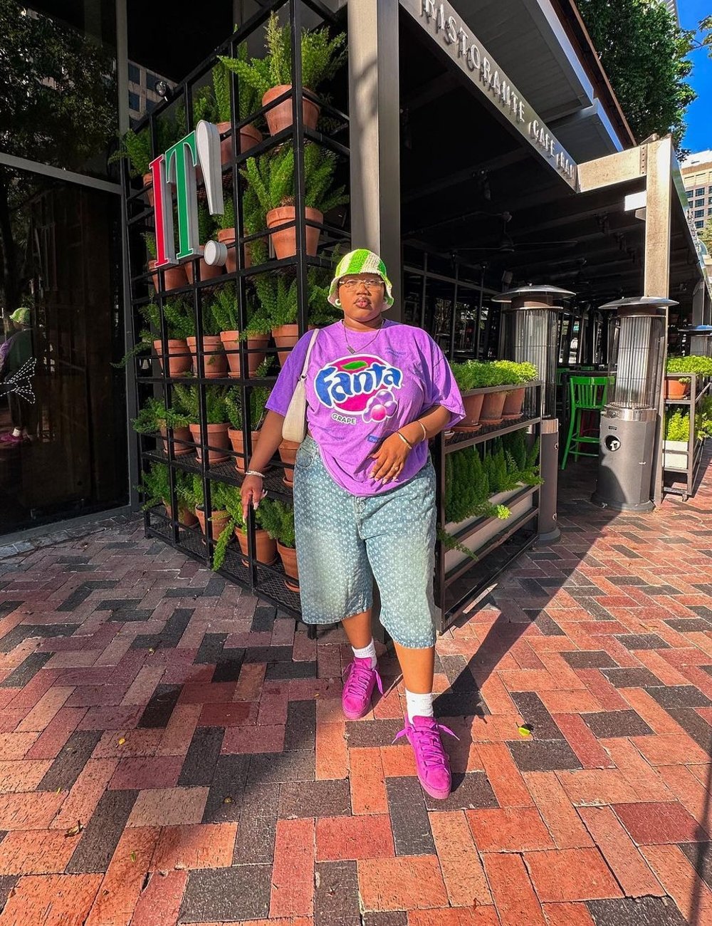 Pessoa vestindo camiseta roxa oversized com estampa da Fanta, bermuda jeans azul clara, tênis roxo, meias brancas e boné verde neon. Estilo casual urbano com foco em cores vibrantes e tendências retrô. Cena capturada em ambiente externo em área urbana.