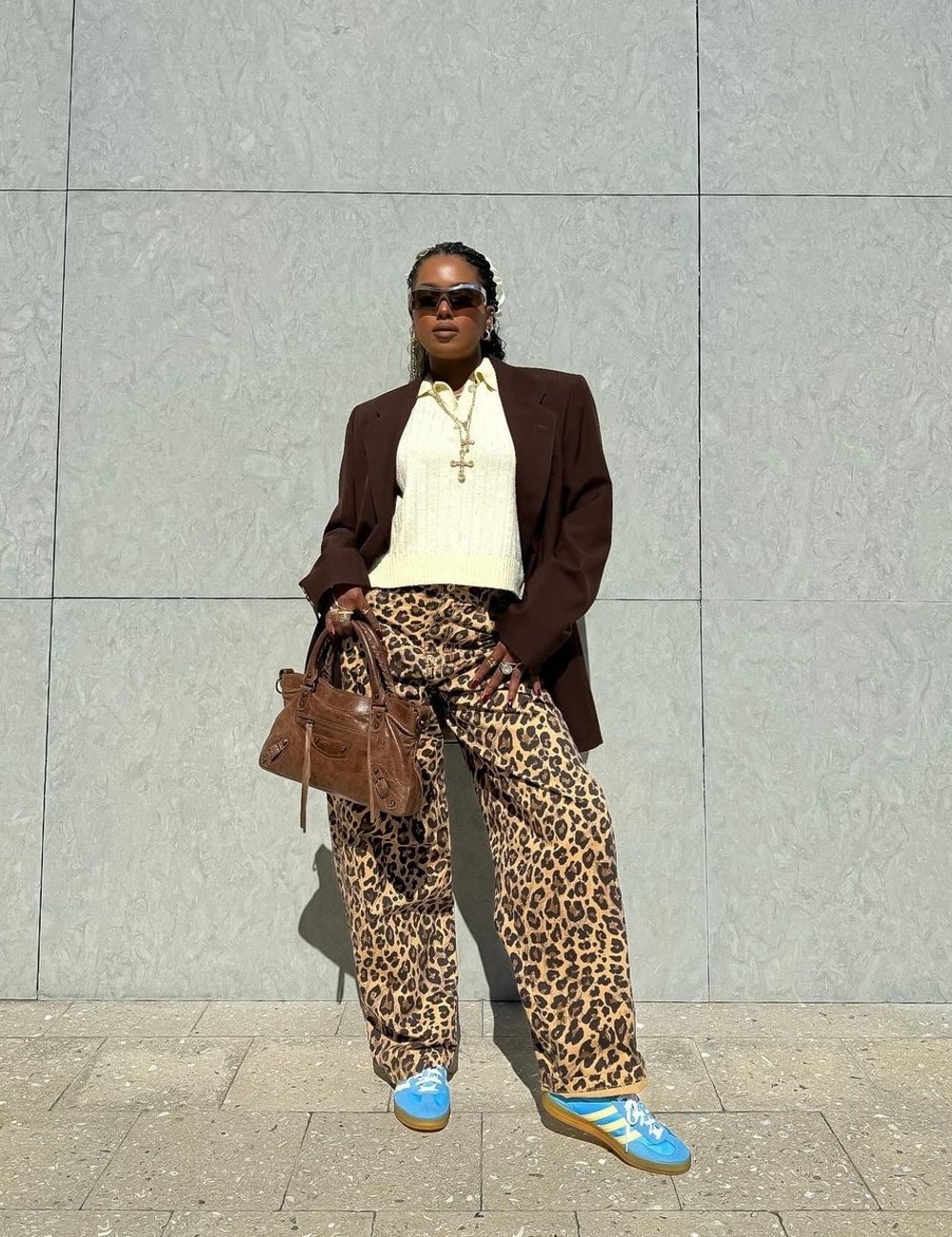 Pessoa vestindo calça com estampa animal print (onça), suéter creme e blazer marrom oversized. Óculos de sol grandes, colar dourado e tênis azuis adicionam toque descolado. Bolsa marrom completa look moderno e eclético.