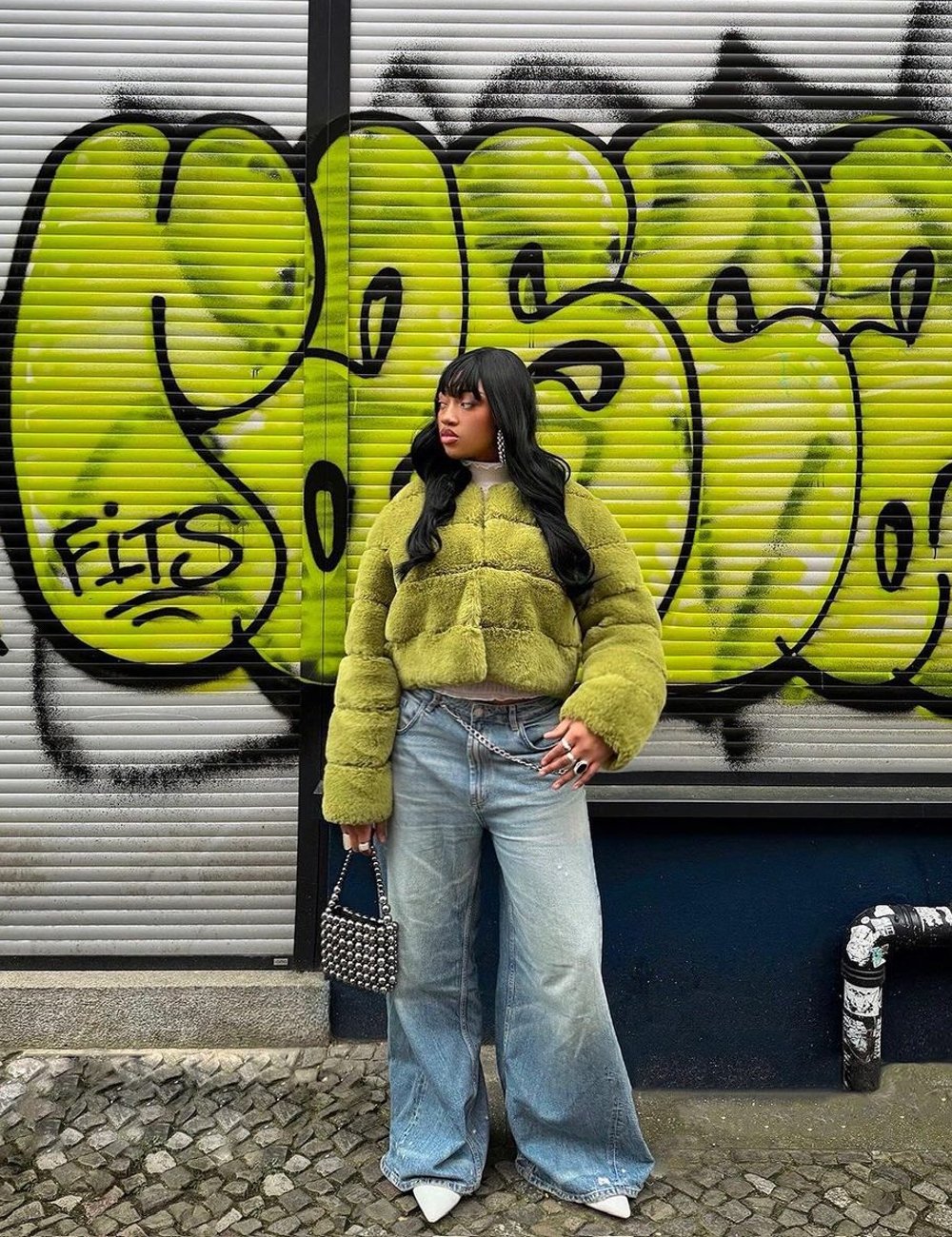 Pessoa vestindo casaco verde peludo e calça jeans oversized estilo anos 90, com corrente na cintura. Complementa o look com bolsa de miçangas e sapatos pontudos brancos. Fundo com grafite colorido, destacando a moda urbana e street style.