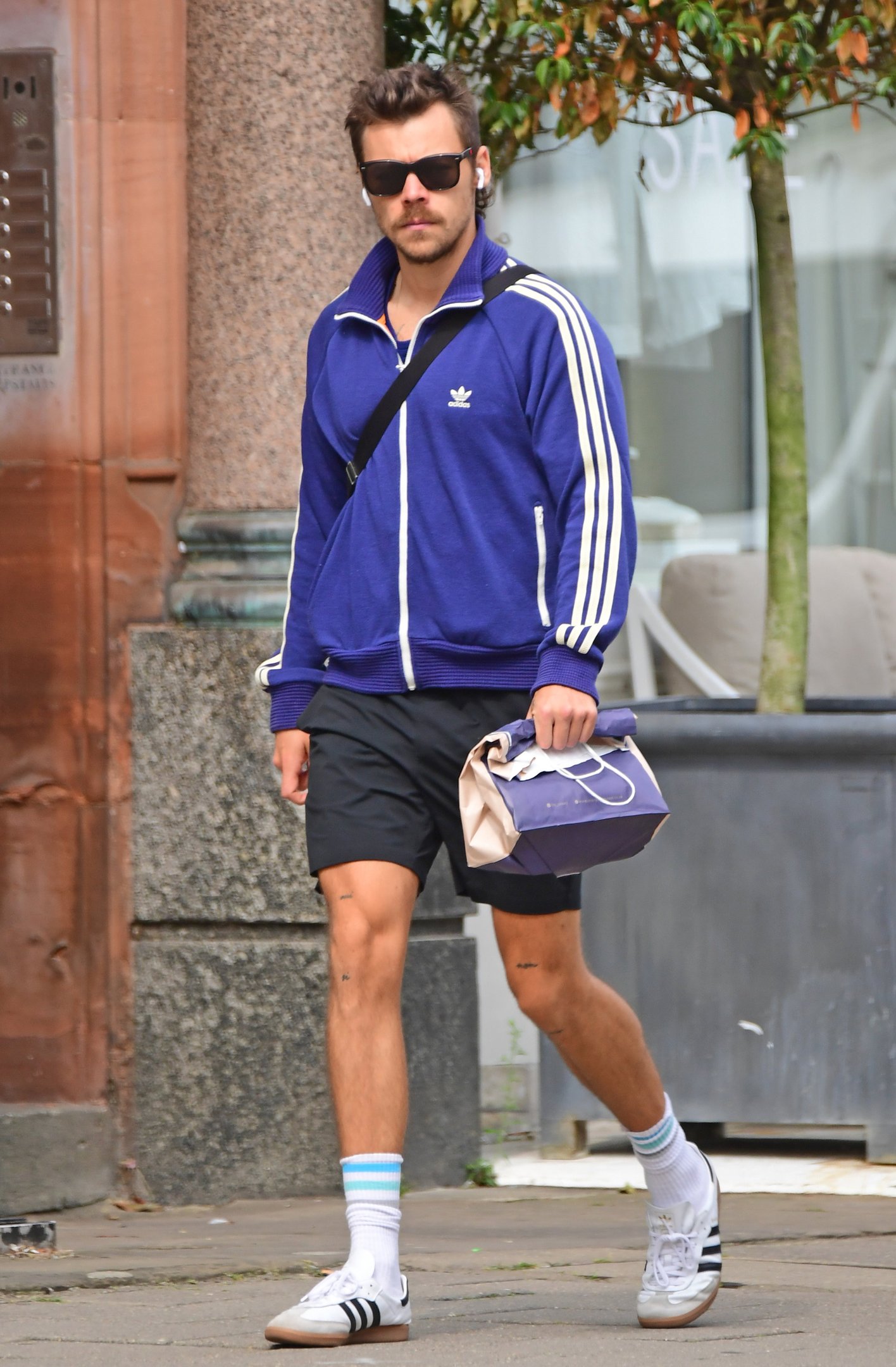 Homem vestindo um agasalho Adidas azul com listras brancas, bermuda preta, tênis branco com listras pretas e meias brancas com detalhe azul. Complementando o look, ele usa óculos escuros e carrega uma bolsa. Estilo casual esportivo.