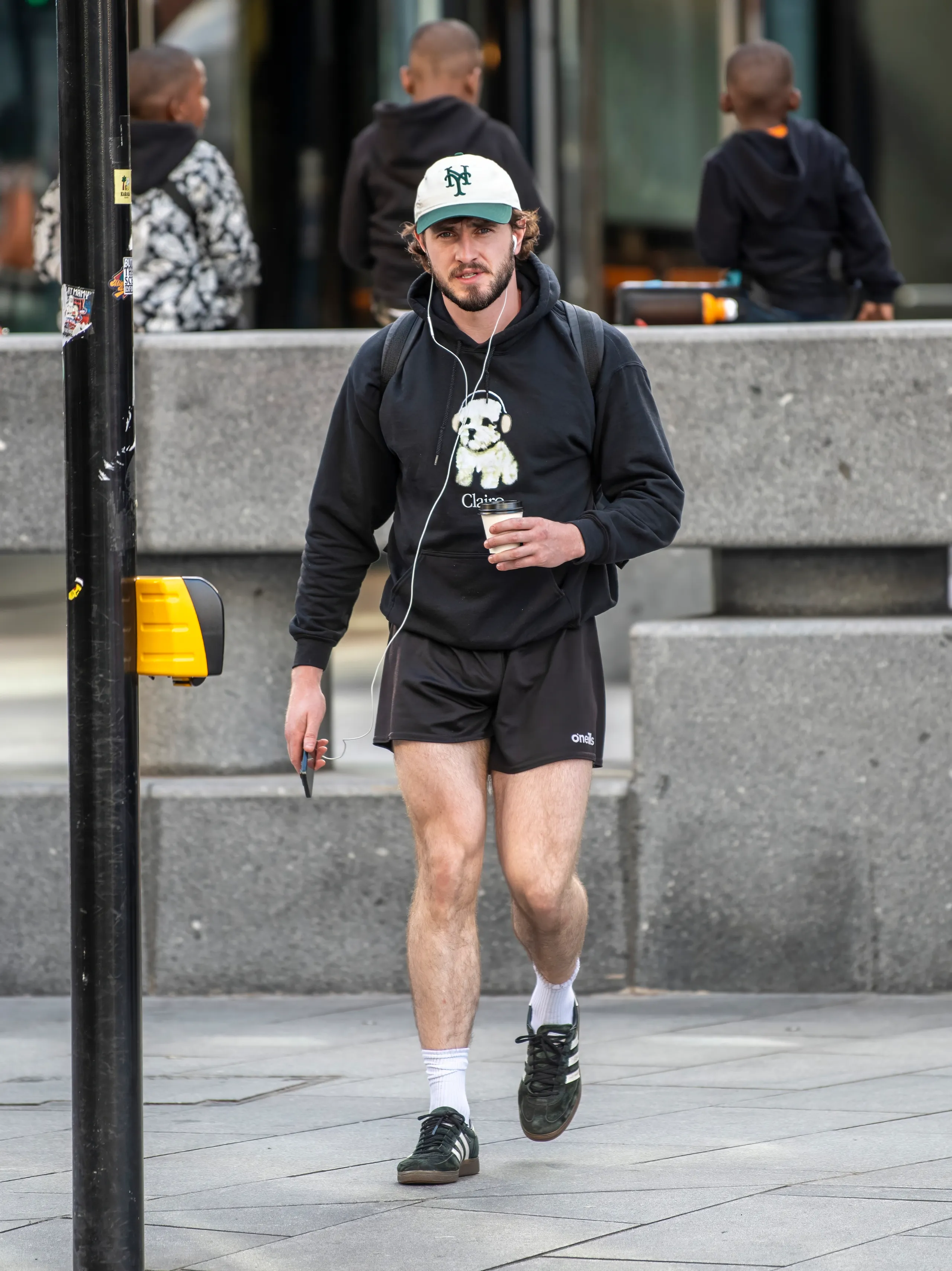 Homem trajando estilo casual esportivo urbano. Ele usa boné branco e verde, moletom preto com estampa de urso, shorts esportivos pretos, tênis preto com branco e meias brancas. Acessórios incluem fones de ouvido e mochila preta.