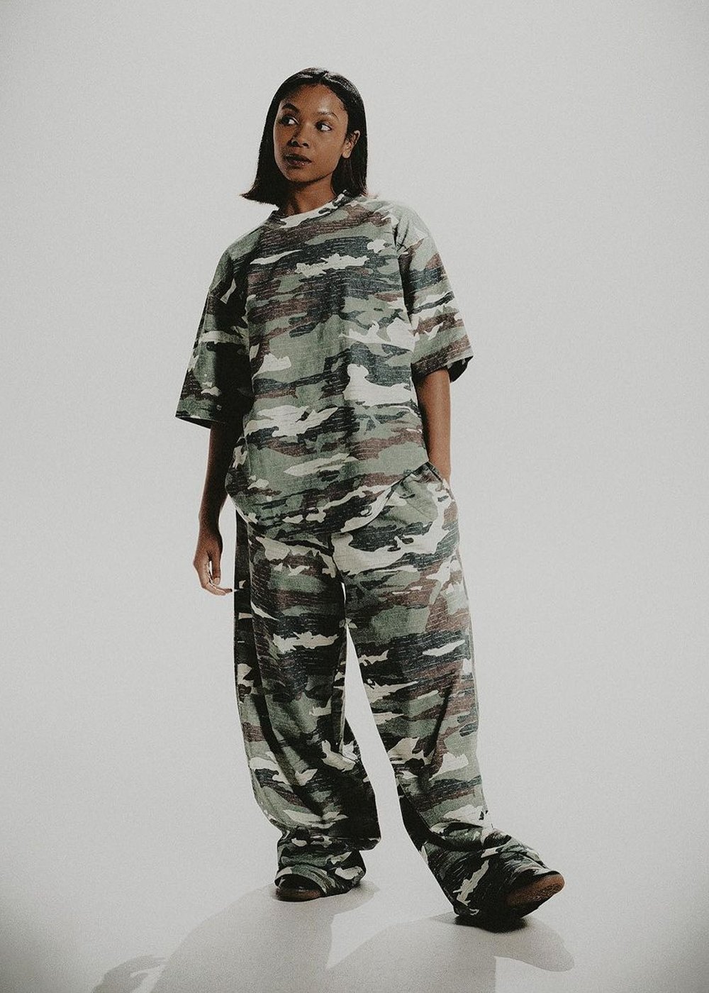 Mulher vestindo conjunto oversized de calça e camiseta com estampa camuflada verde. Tendência de moda streetwear com inspiração militar, destacando conforto e estilo urbano.