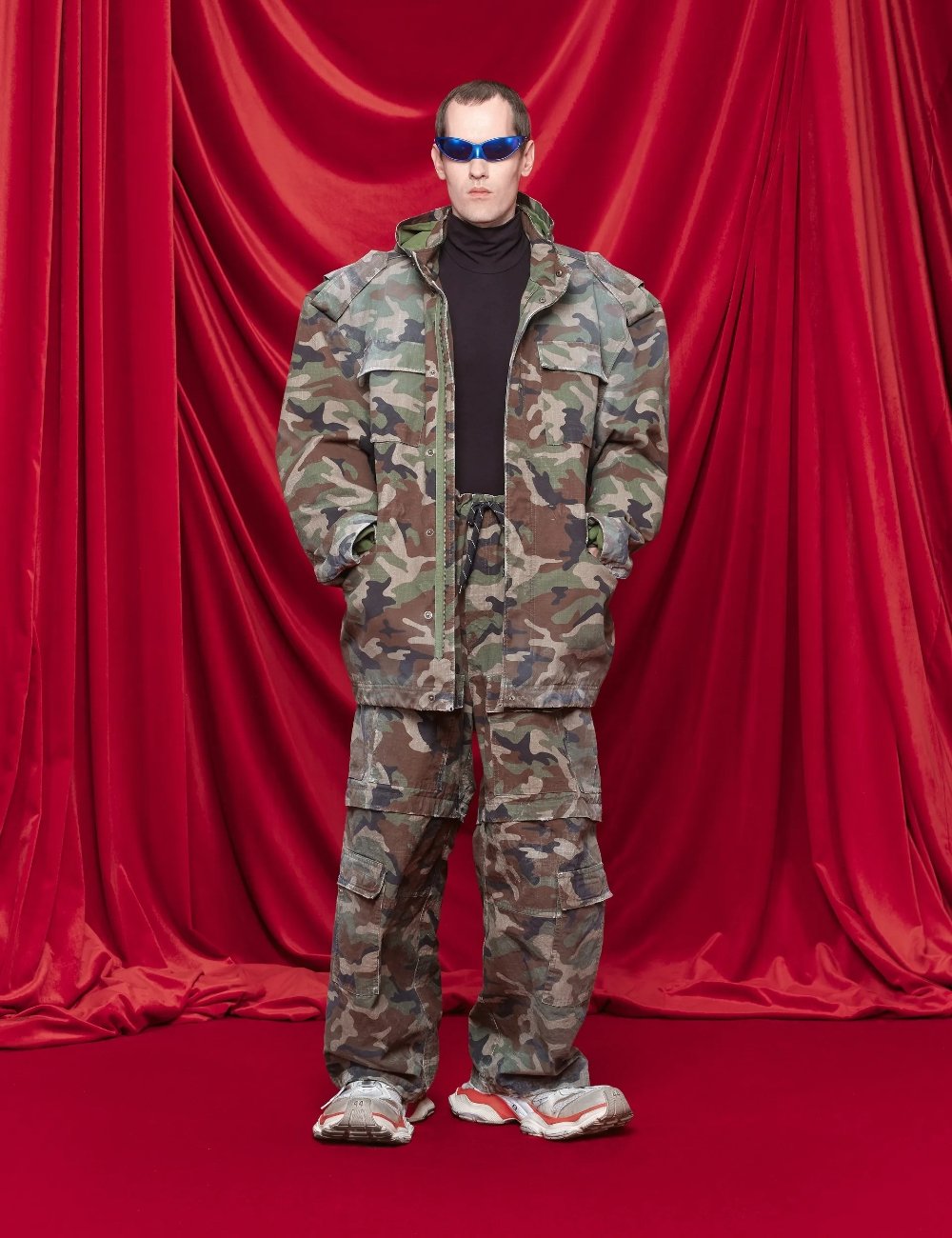 Homem vestindo jaqueta de estampa camuflada, calças cargo no mesmo padrão, suéter preto de gola alta e tênis volumosos. Completa o look com óculos escuros futuristas azuis. Estilo utilitário e militar combinado com tendências modernas de moda urbana.