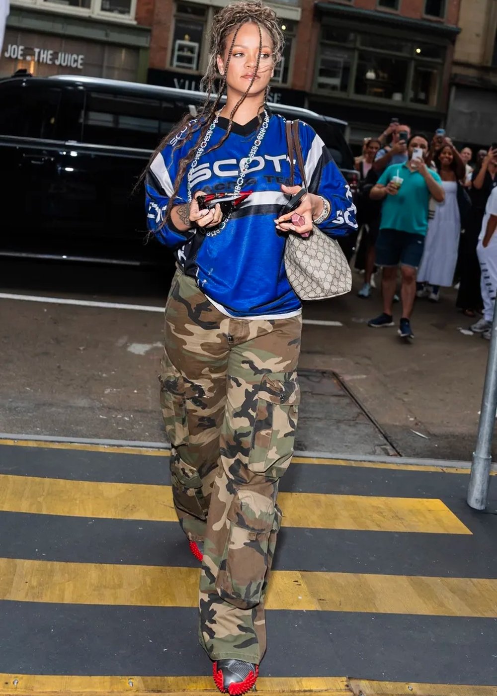 Rihann usando uma camisa de times esportivos azul e calças cargo de estampa camuflada. A moda é influenciada pelo estilo streetwear, com uma mistura de peças esportivas e utilitárias. Destacam-se também acessórios como colar longo e bolsa de ombro.