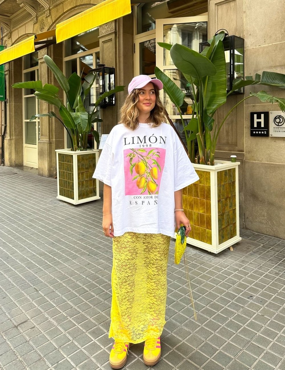 Jovem vestindo camiseta oversized com estampa de fruta, saia de renda amarela sobre saia curta e tênis amarelos com listras rosas. O look é complementado por boné rosa e bolsa verde com corrente dourada.