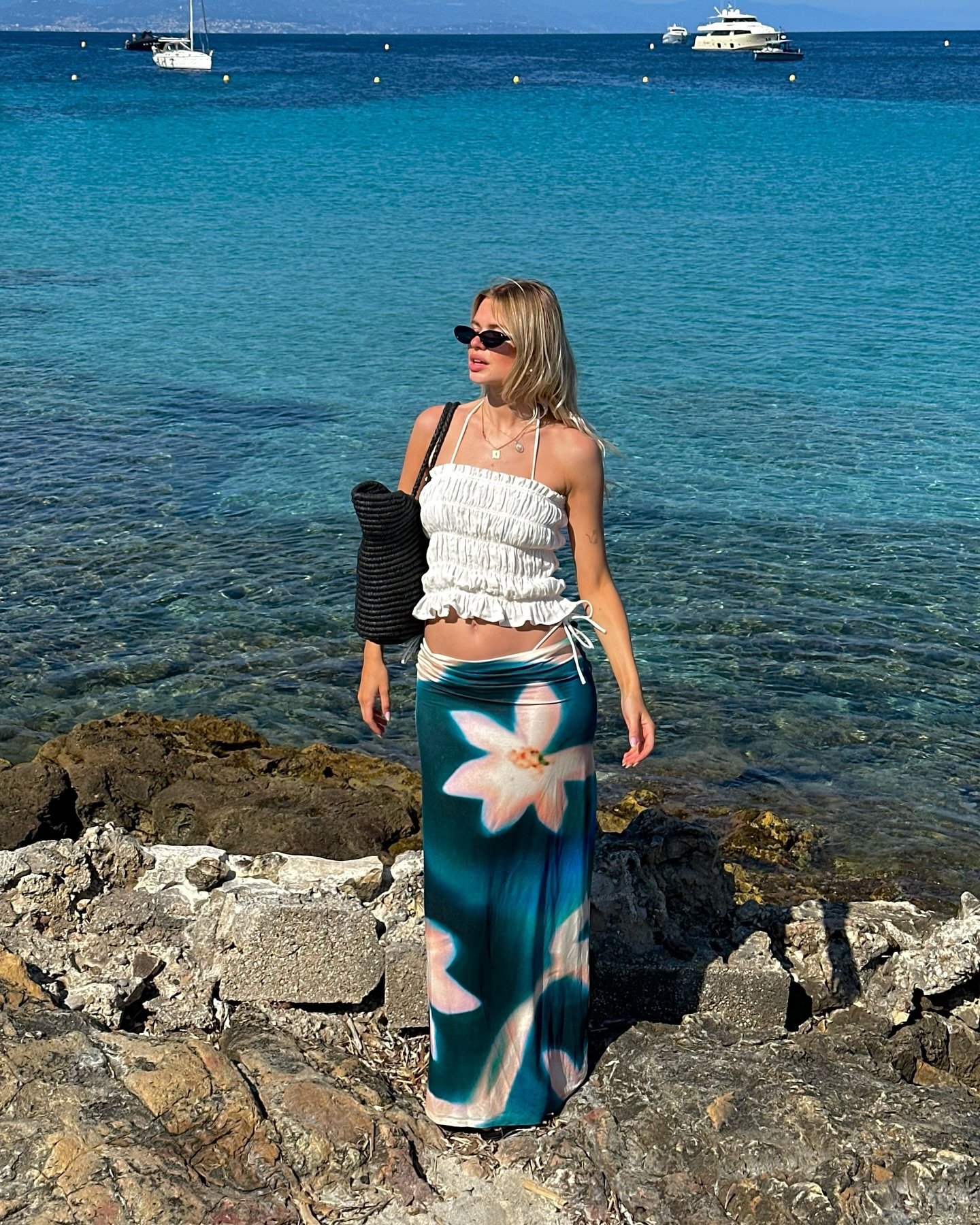 Mulher vestida com top branco franzido e saia longa azul com estampa floral, adicionando um toque veranil e elegante ao look. Ela complementa com óculos escuros e bolsa preta. Paisagem de praia e clima tropical ao fundo realçam o estilo boho-chic.