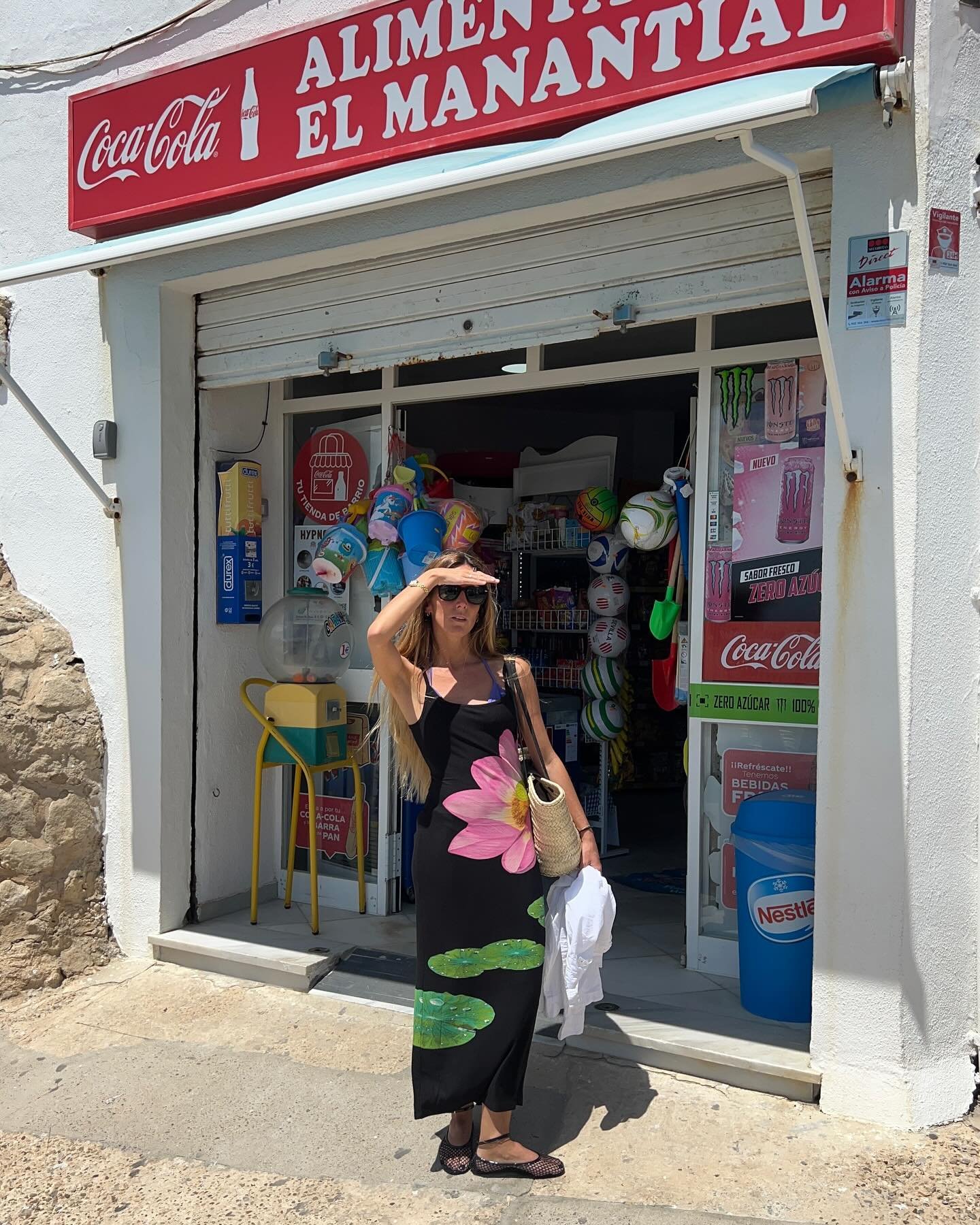 Mulher usa vestido preto longo com estampa de flor rosa, óculos de sol e bolsa de palha em frente a uma loja de conveniência 