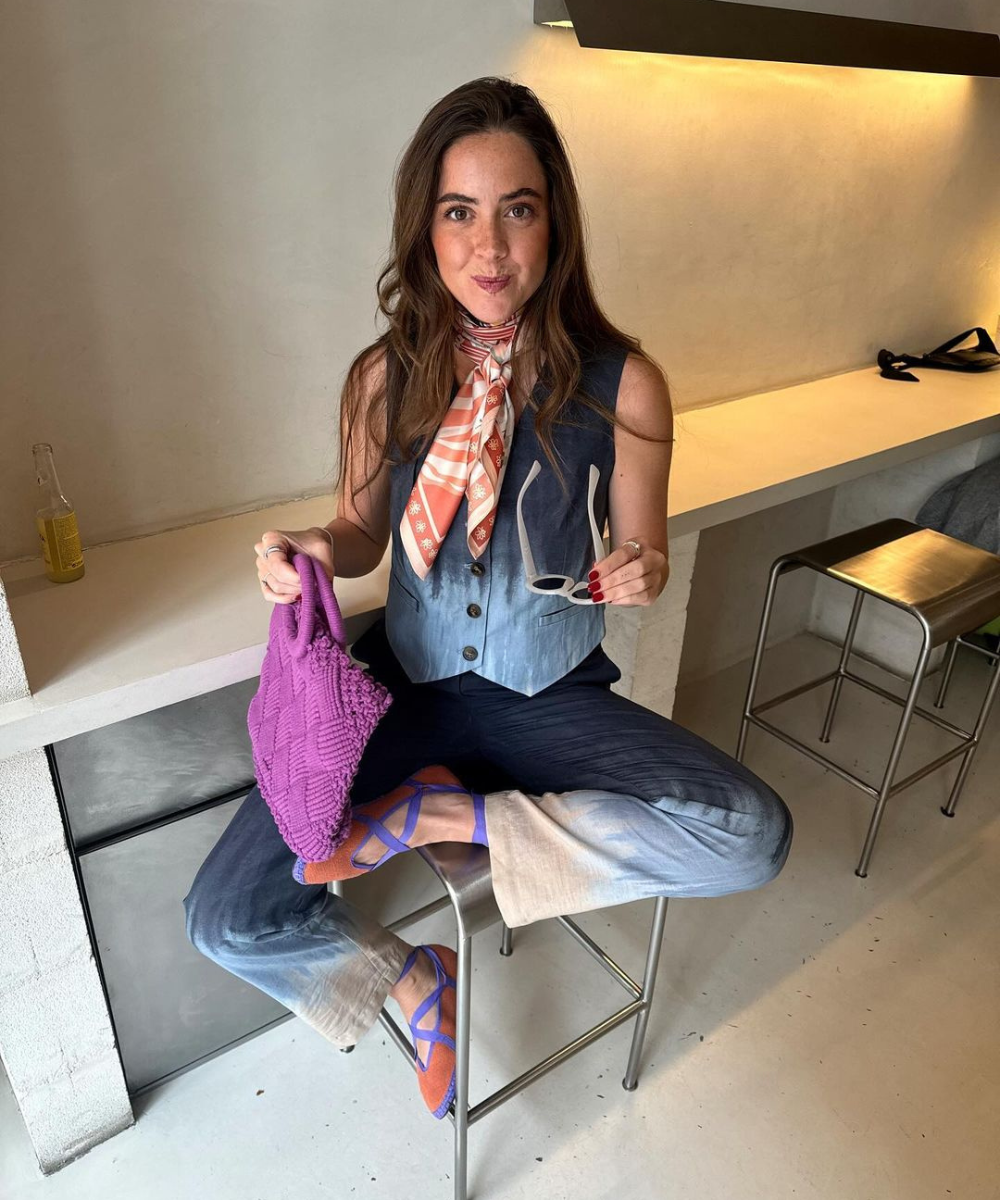 Mulher vestindo colete jeans e calça de lavagem dégradé, lenço laranja estampado no pescoço, sapatos estilosos laranja com detalhes roxos e bolsa de tricô roxa. Carrega óculos de sol brancos. Visual despojado e moderno.