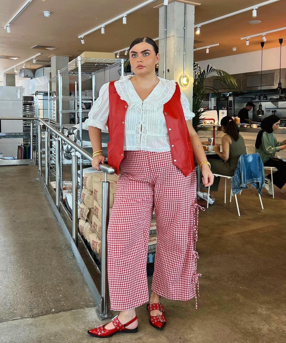 Mulher vestindo blusa branca rendada com babados, colete vermelho em material brilhoso, calça xadrez vermelha de fundo branco com amarrações laterais e sapatos vermelhos com detalhes vazados. Look combina tendências vintage e modernas.