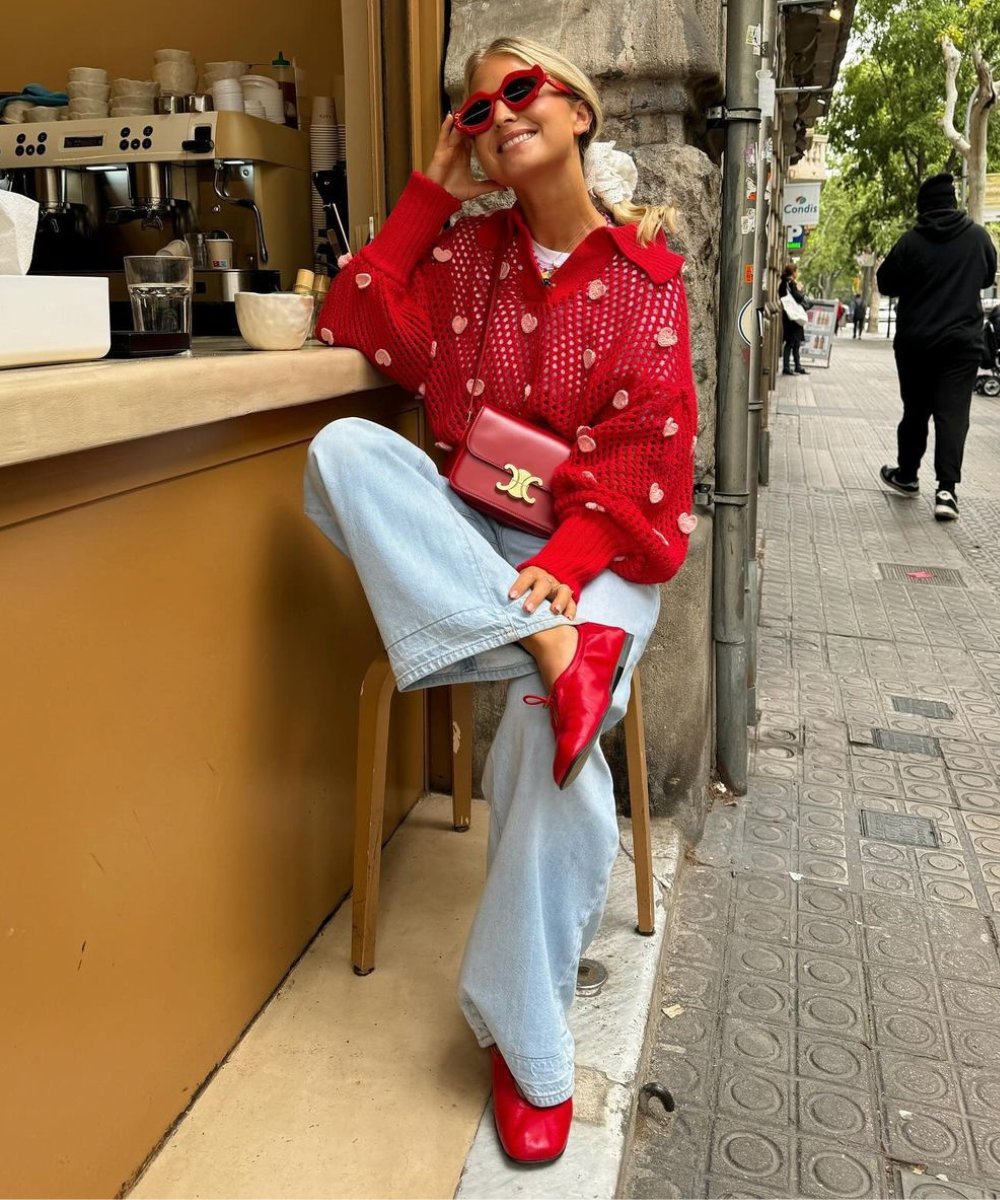 Mulher com suéter vermelho oversized, bordado com detalhes em corações, calças jeans largas e sapatos vermelhos combinando com a bolsa de mão e óculos de sol. Look moderno e casual combinando tons de vermelho e jeans, ideal para um dia descontraído na cidade.