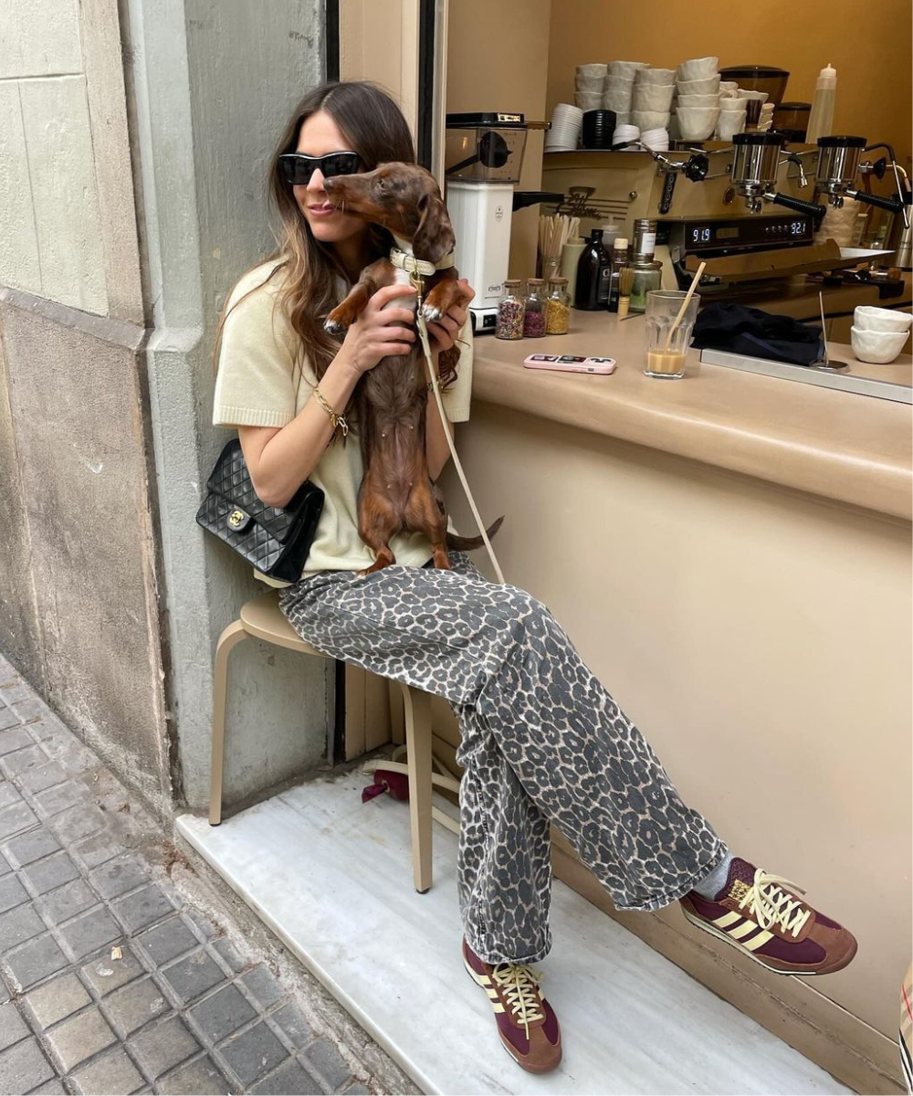 Mulher com óculos escuros e bolsa preta de grife, segurando um cachorro. Ela usa blusa bege, calça com estampa animal print e tênis adidas marrom/vermelho. Estilo casual e moderno.