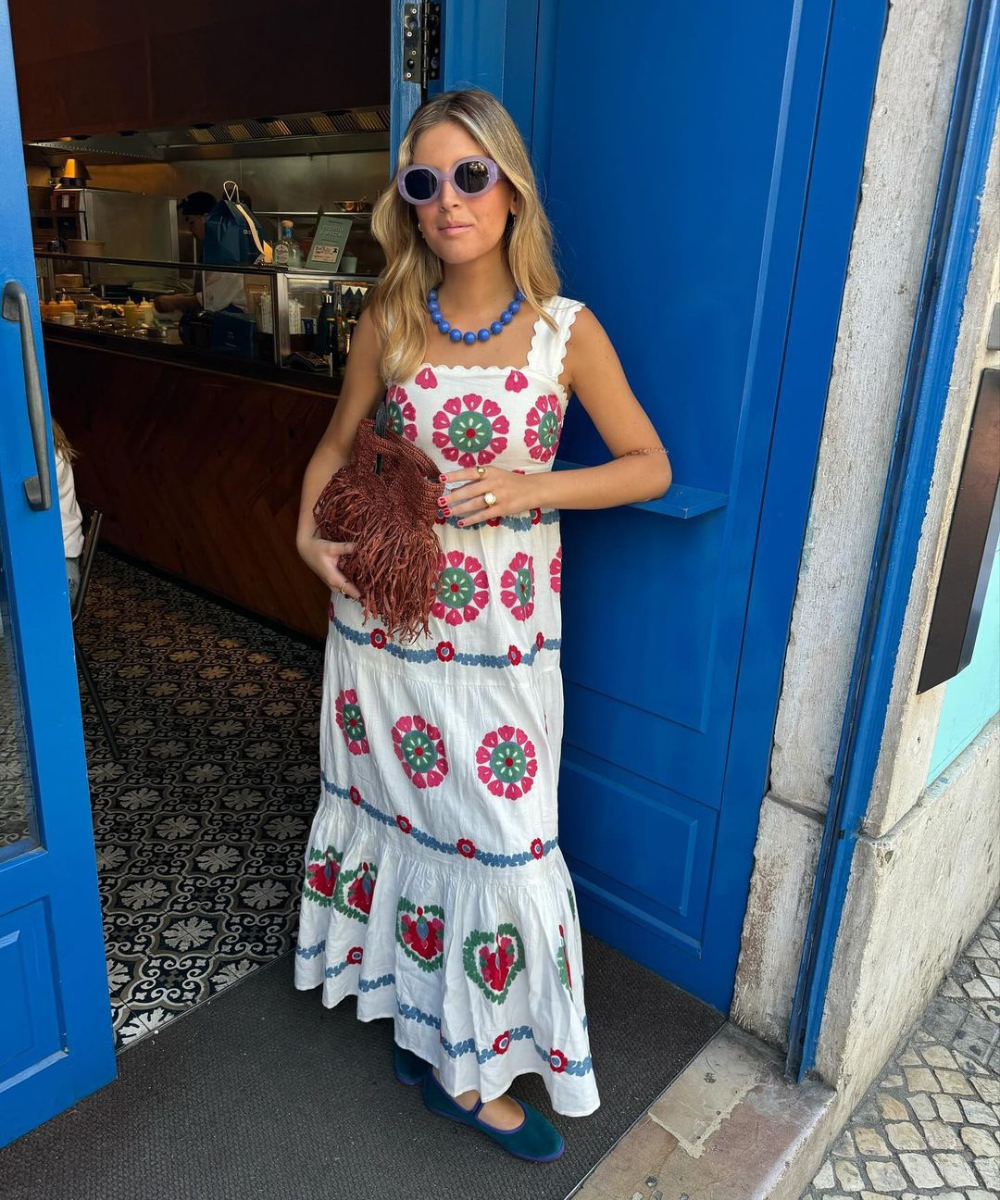 Mulher vestindo um vestido longo branco com estampas florais vermelhas e verdes, colar de contas azuis e óculos de sol redondos. Ela segura uma bolsa marrom de franjas. O estilo é boho chic com toques vintage.