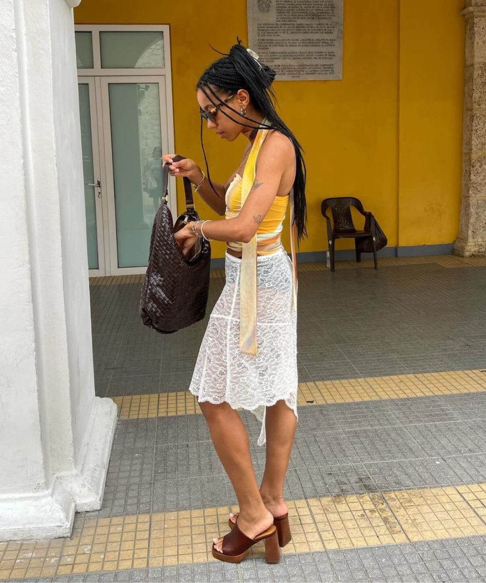 Mulher com tranças longas e óculos de sol, vestindo top amarelo, saia rendada branca e sandálias de salto alto marrom. Carrega uma bolsa preta grande. Look de verão descontraído e elegante, com mistura de texturas e acessórios minimalistas.