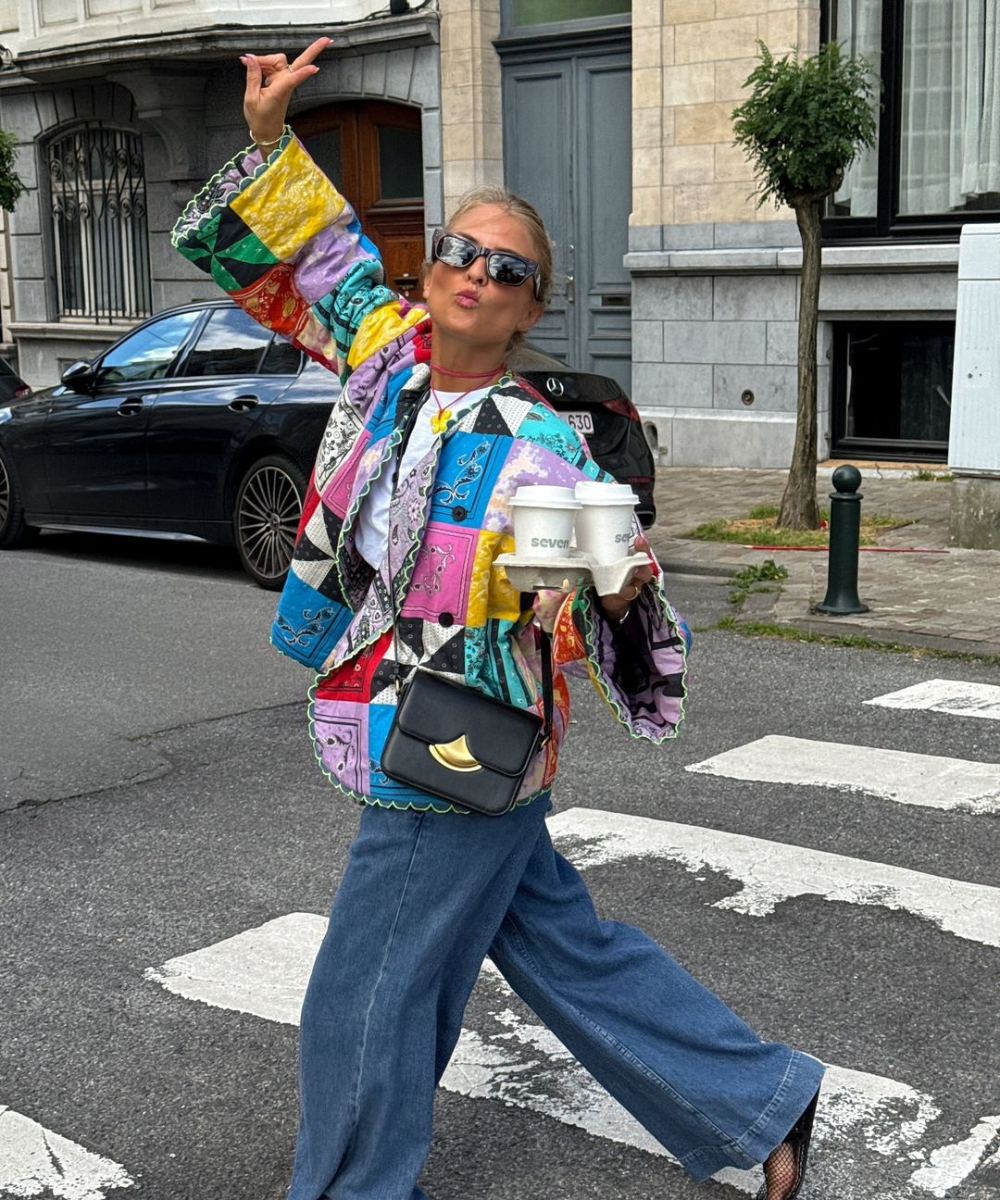 Pessoa vestindo casaco de crochê colorido com estampas florais, saia longa preta com detalhes recortados e sandálias de tiras em um ambiente casual e aconchegante. Tendências: moda artesanal, crochê, estampas florais, eclético vintage, boho-chic.