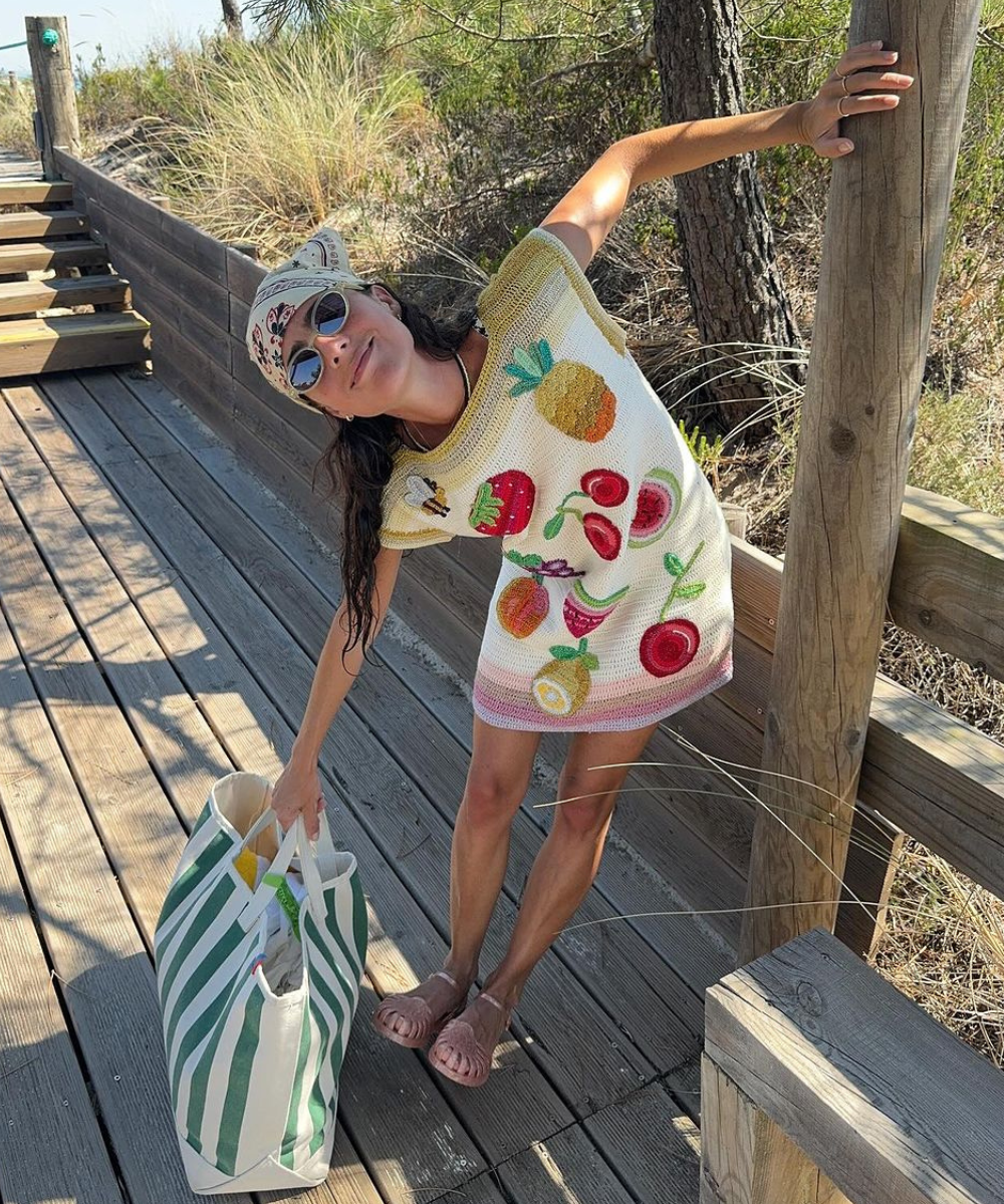 Pessoa usando vestido de crochê com estampas de frutas, óculos escuros, chapéu estilo bandana e sandálias rasteiras. Carrega uma bolsa grande com listras verdes e brancas. Estilo casual e alegre, ideal para um dia de verão.