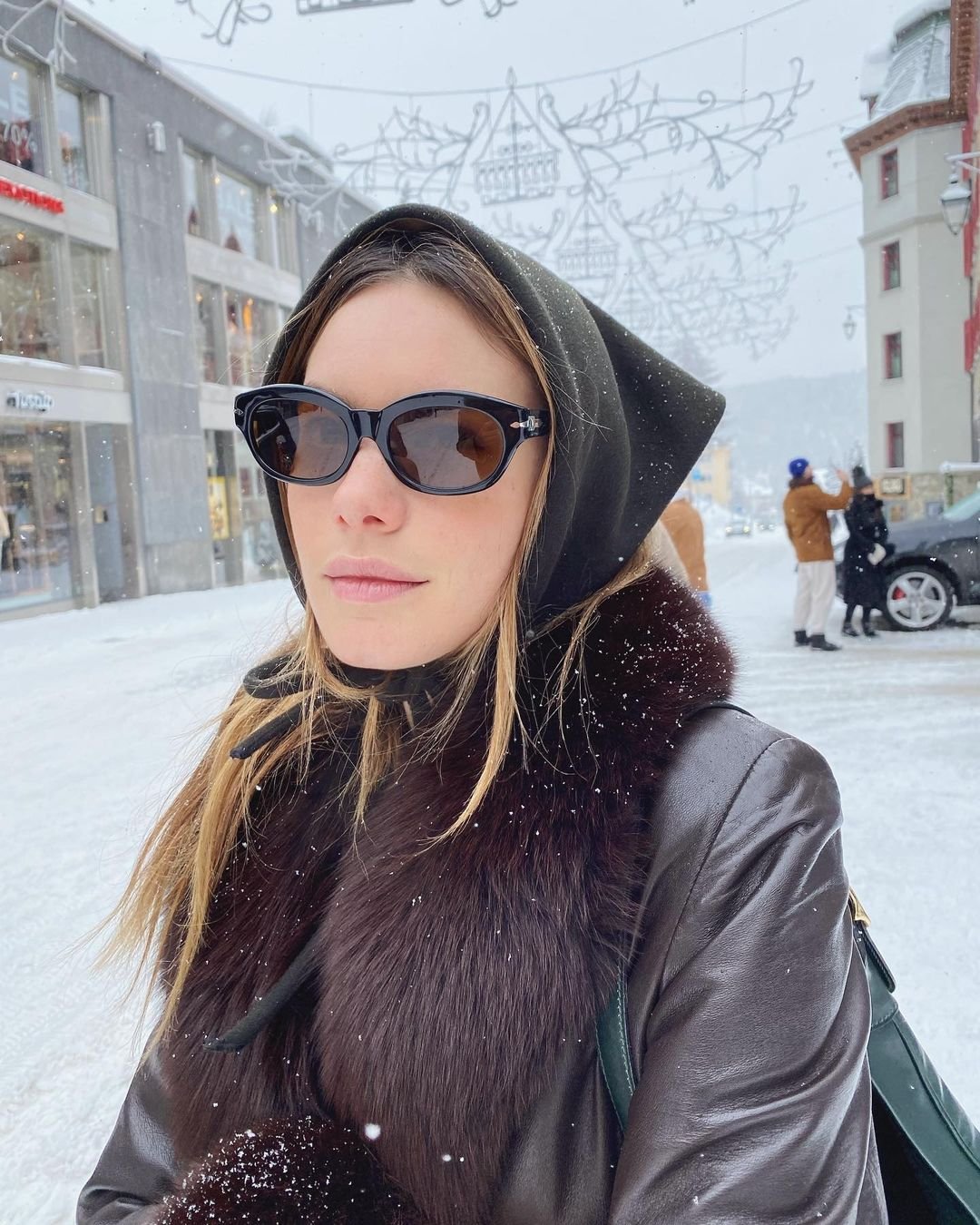 Mulher com óculos de sol, usando capuz preto e casaco de couro com gola de pele marrom, no cenário de uma rua nevada. Look sofisticado e elegante, destacando tendências de moda inverno com foco em acessórios e materiais quentes e luxuosos.