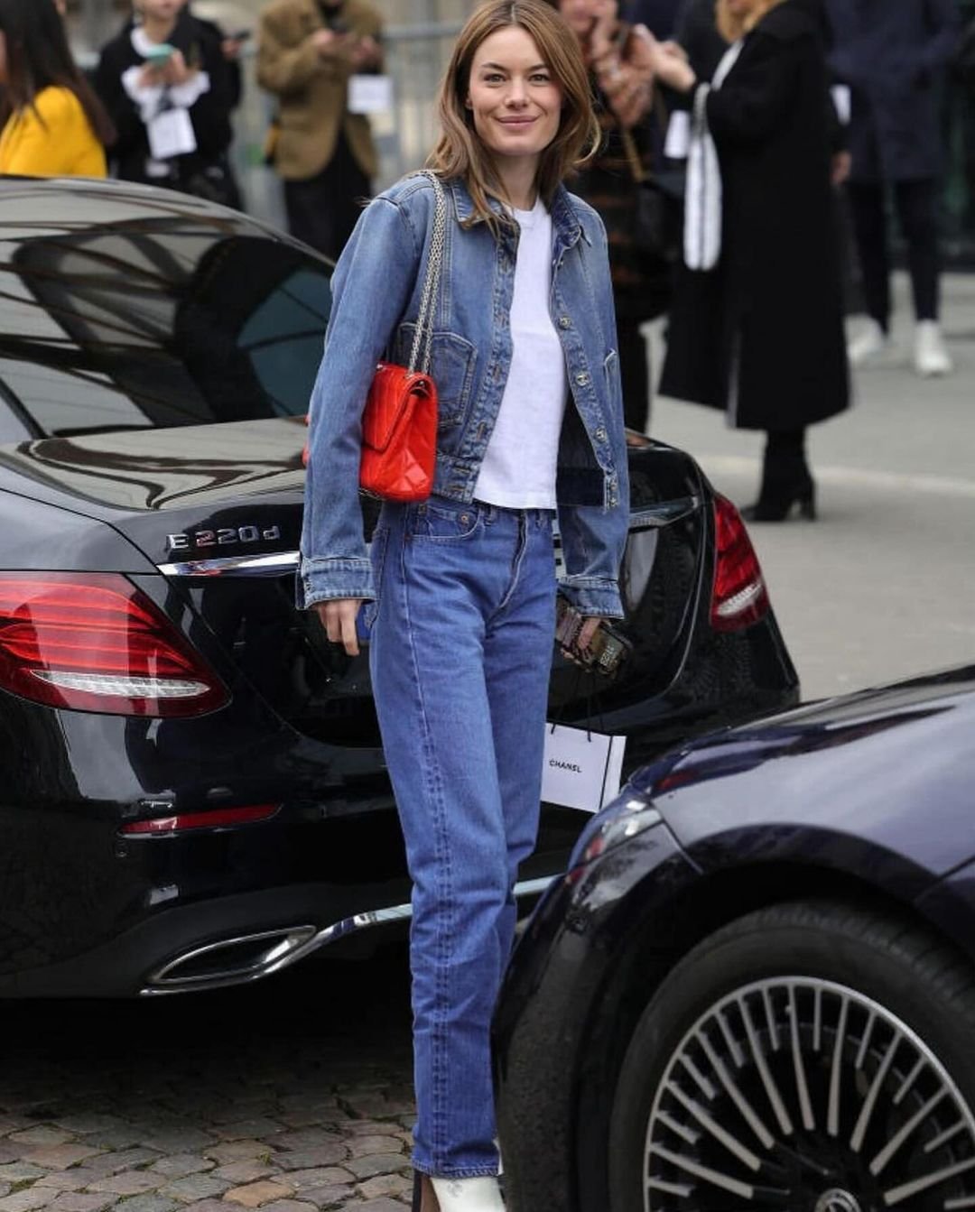 Mulher com visual casual-chic, usando jaqueta jeans, camiseta branca, calça jeans de cintura alta e botas brancas. A bolsa vermelha destaca-se como acessório de cor vibrante. O look reflete tendências de moda urbana e minimalista.