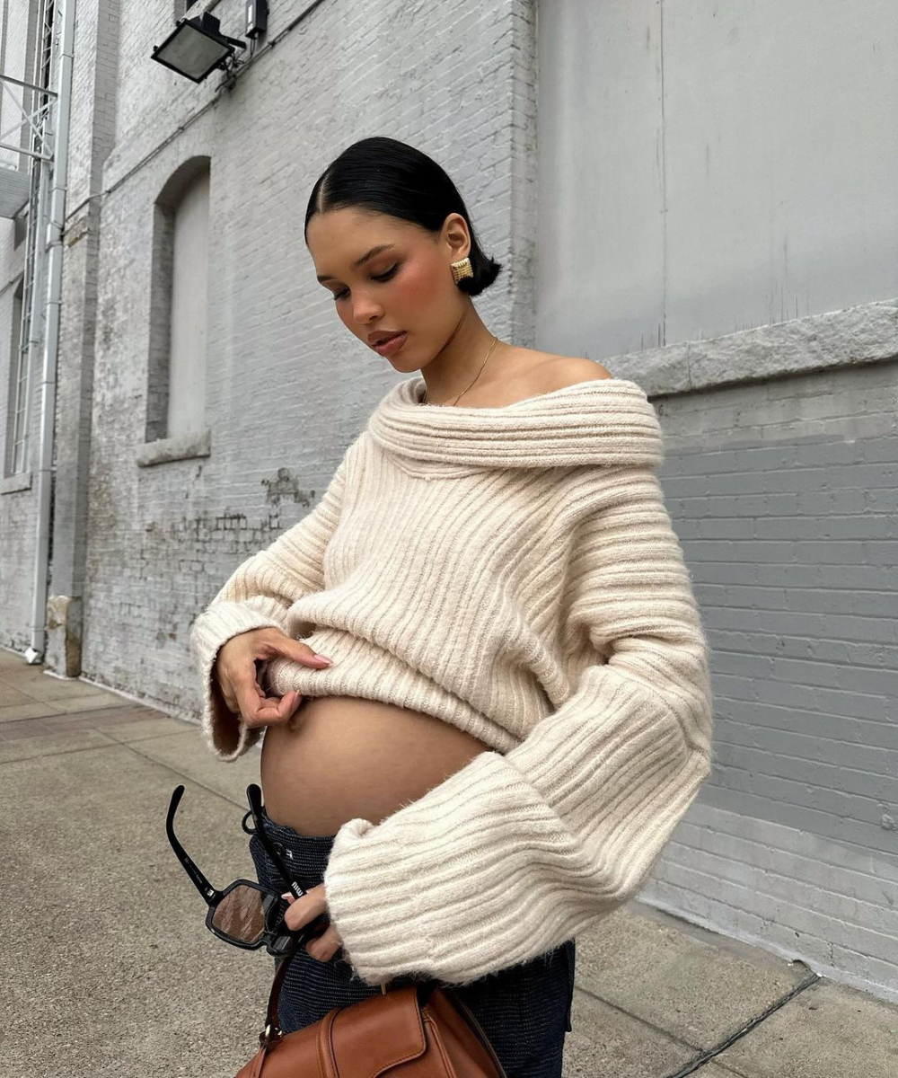 Mulher grávida usando suéter oversized de tricô creme com ombro caído, cabelo preso, brincos dourados grandes e calça escura. Tendências: moda confortável, peças de tricô, acessórios statement e tons neutros. Segurando uma bolsa marrom e óculos de sol.