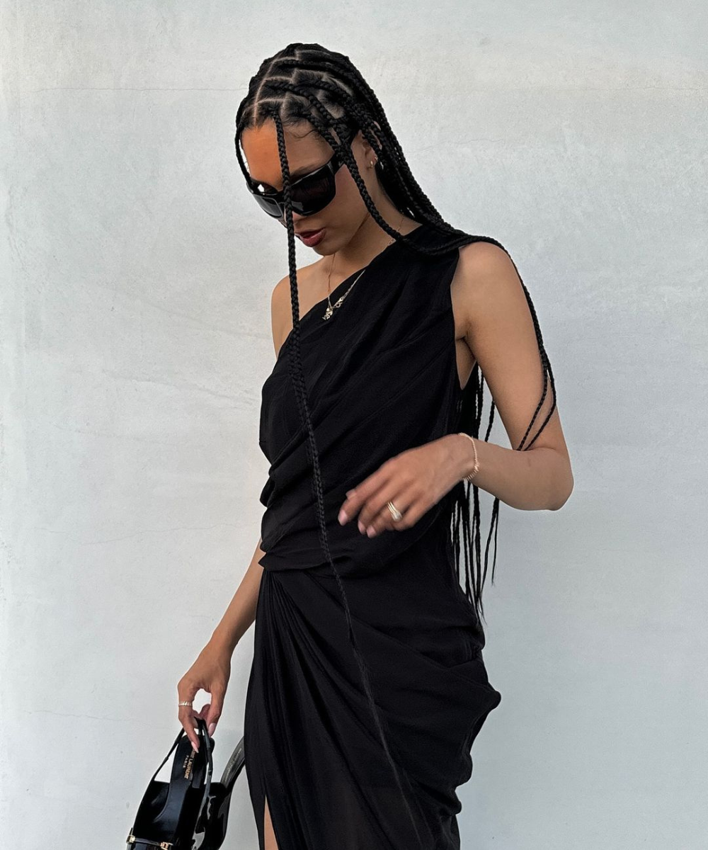 Mulher usando vestido preto de um ombro só, óculos de sol grandes e cabelo com tranças box braids. Acompanha bolsa preta pequena. Look moderno e sofisticado, destacando tendências de moda minimalista e elegante.