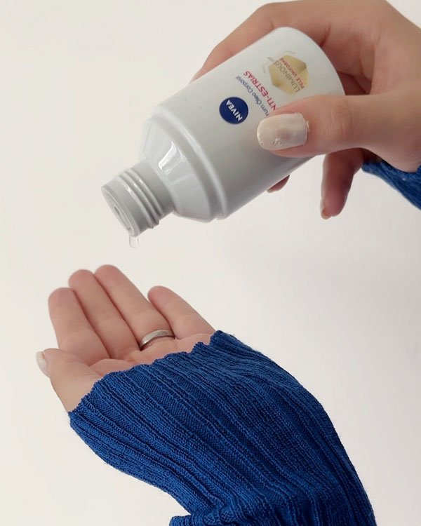 Pessoa em suéter azul despeja óleo corporal da marca Nivea White Crise nas mãos. Unhas com esmalte branco e suéter de malha azul são destaques, refletindo tendência de roupas confortáveis e autocuidado.