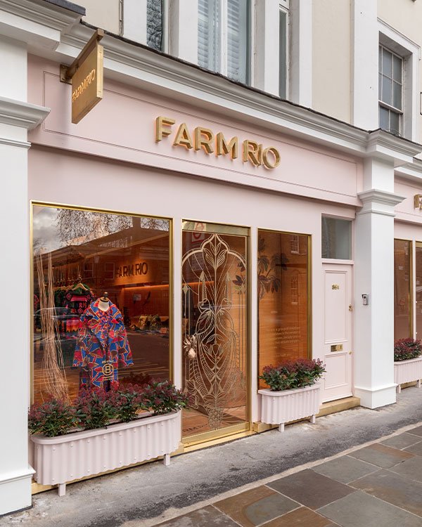 Fachada da loja FARM Rio, com visual moderno em tons de rosa e dourado. Vitrine destaca vestido vibrante com estampas tropicais coloridas, tendência da moda brasileira. Decoração externa com plantas em vasos brancos. Design sofisticado e atraente.