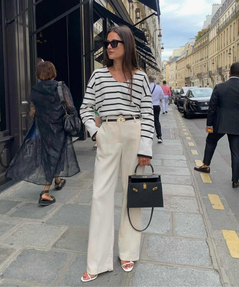 Mulher usando blusa listrada branco e preto com calça de cintura alta e pernas largas em tom de creme. Complementa o visual com óculos de sol, bolsa de mão preta e sandálias brancas de salto baixo. Estilo elegante e casual, ideal para street style mo