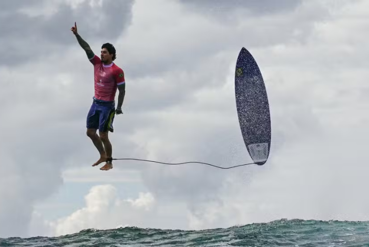 Surfista com camiseta rosa e bermuda azul realiza uma manobra com prancha no ar. Foto histórica das Olímpiadas de Paris.