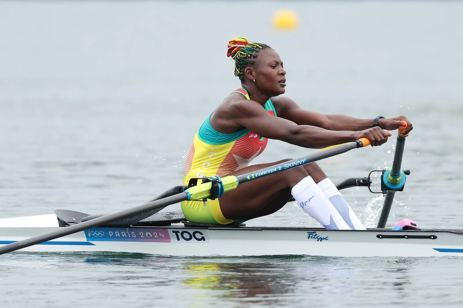 Atleta remando com uniforme colorido nas cores verde, amarelo e vermelho, e meias brancas de compressão. Cabelos com tranças multicoloridas, tendência vibrante e esportiva. Detalhe do barco com inscrições 