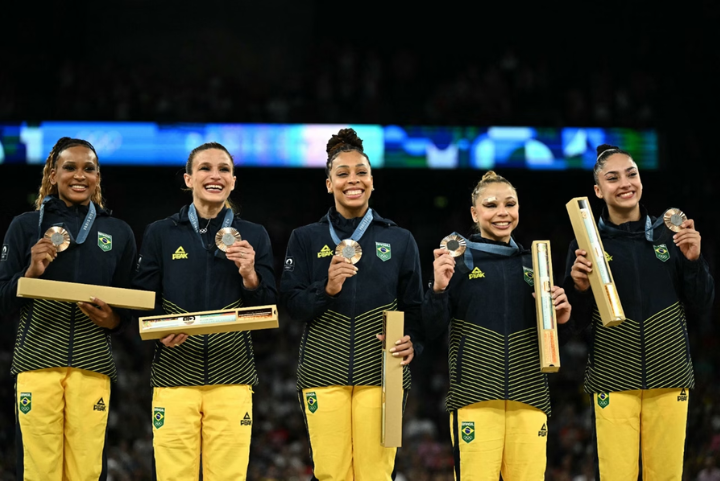 Atletas brasileiras exibem medalhas e troféus enquanto vestem uniformes esportivos estilizados, compostos por jaquetas azuis marinho com detalhes em verde e amarelo e calças amarelas. O conjunto alia conforto e estilo, destacando as cores nacionais.