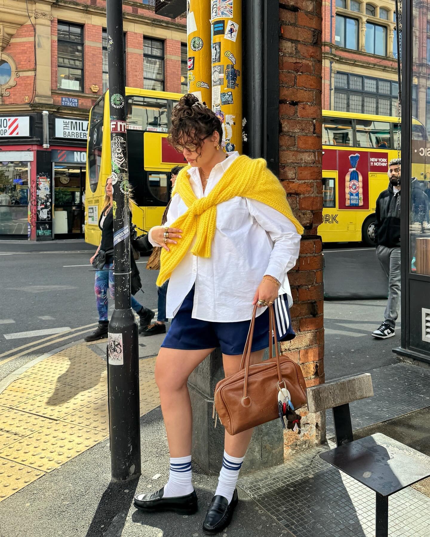 Roupas casuais de rua: camisa branca oversized, shorts azul-marinho, suéter amarelo sobre os ombros, meias brancas com listras azuis e mocassins pretos. Acessórios incluem óculos de sol e bolsa de couro marrom. Estilo urbano e despojado.