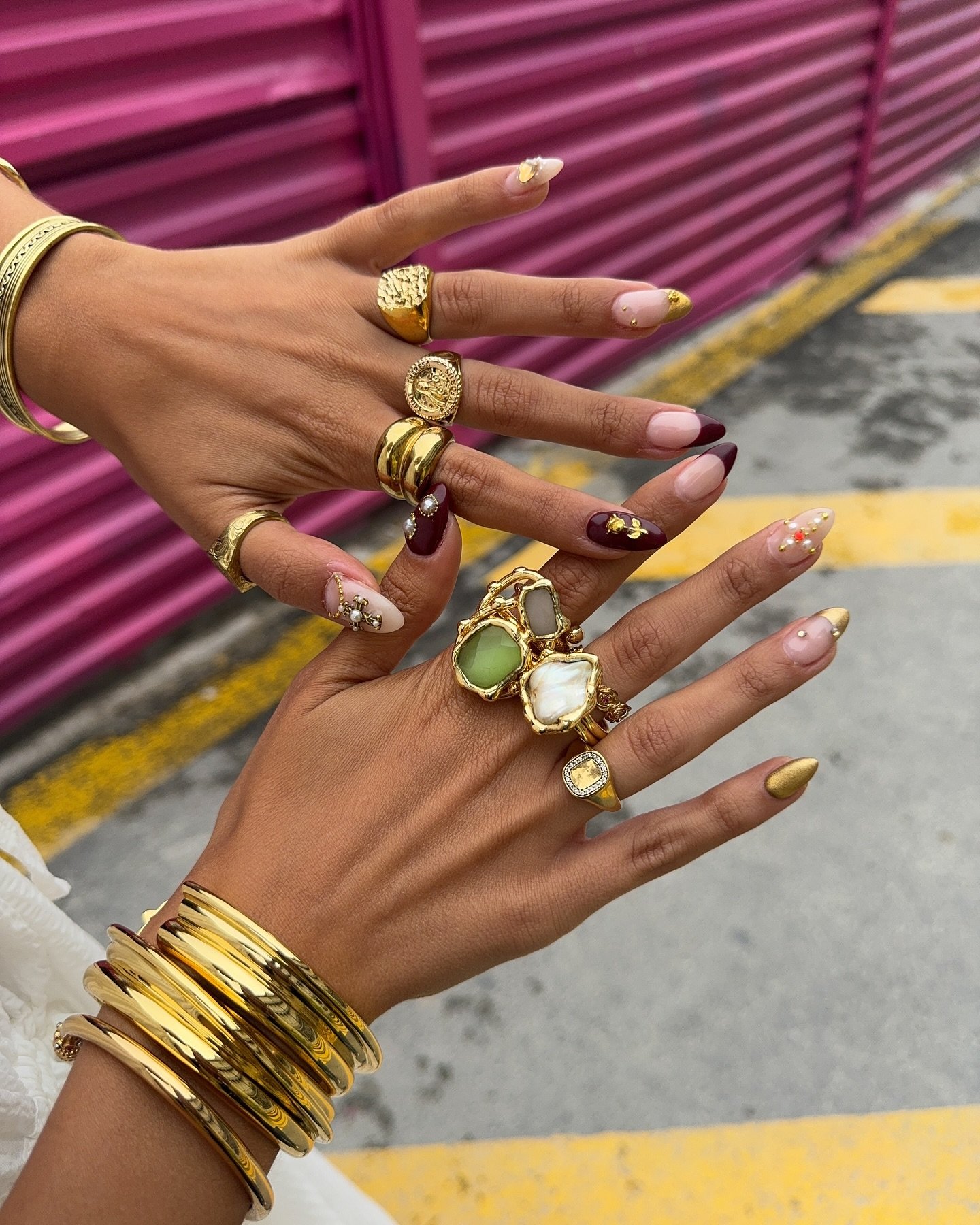 Mãos adornadas com anéis chamativos e pulseiras largas douradas. Unhas longas e decoradas com cores variadas, incluindo vinho, nude e detalhes em dourado. Tendência de moda focada em acessórios exuberantes e nail art sofisticada.