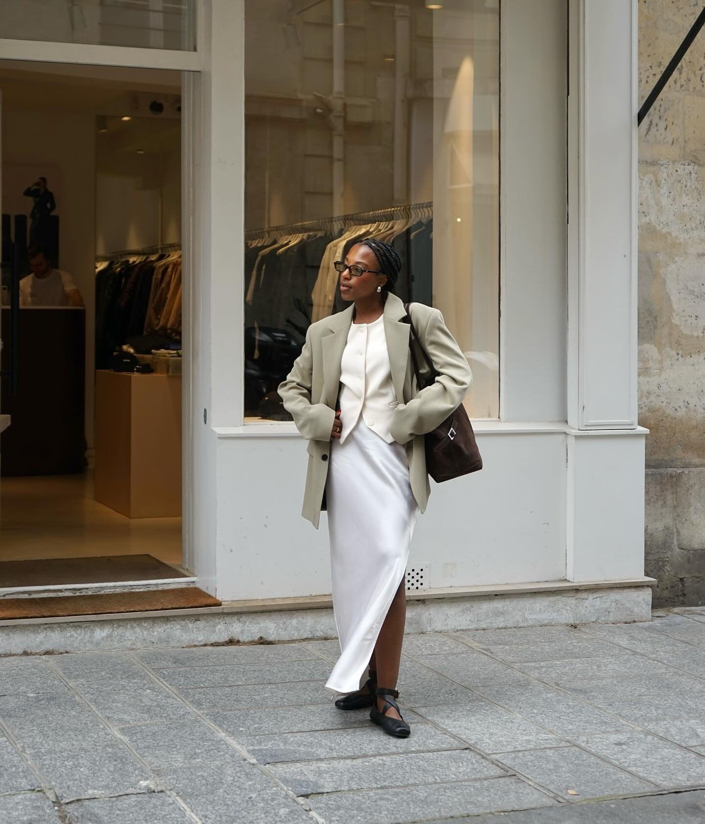 Pessoa bem vestida com look elegante e moderno: blazer oversized verde claro, vestido longo acetinado branco, bolsa marrom e sapatilhas pretas. Óculos de armação escura, cabelo trançado e estilo minimalista. Cenário urbano com loja de roupas ao fundo.