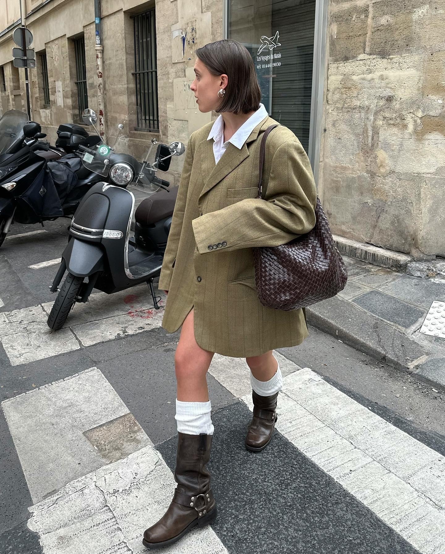 Mulher com visual urbano-chique. Usa blazer oversized bege sobre camisa branca, meia até o joelho combinada com botas marrons e bolsa de ombro texturizada. Estilo moderno e despojado, misturando elementos clássicos e contemporâneos.