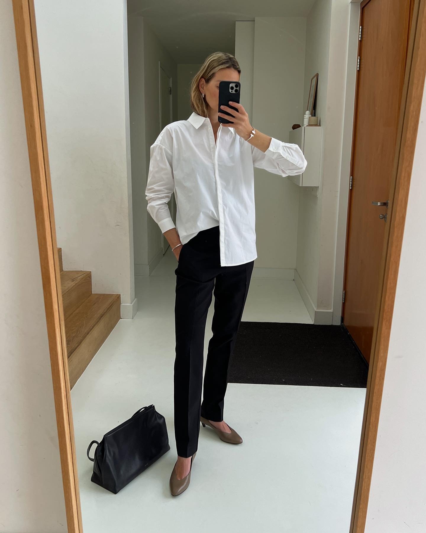 Look minimalista com camisa branca oversized e calça preta de alfaiataria. Bolsa preta estruturada e sapatos de couro marrom. Visual elegante e atemporal, perfeito para ambiente de trabalho ou ocasiões formais. Tendência: simplicidade e elegância.