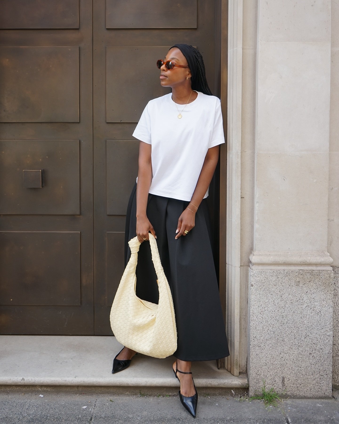 Imagem de moda urbana minimalista: mulher vestindo camiseta branca básica, saia midi preta, sapatos pretos de bico fino e óculos de sol clássicos. A bolsa amarela grande adiciona um toque de cor. Look elegante e moderno, ideal para o cotidiano na cidade.