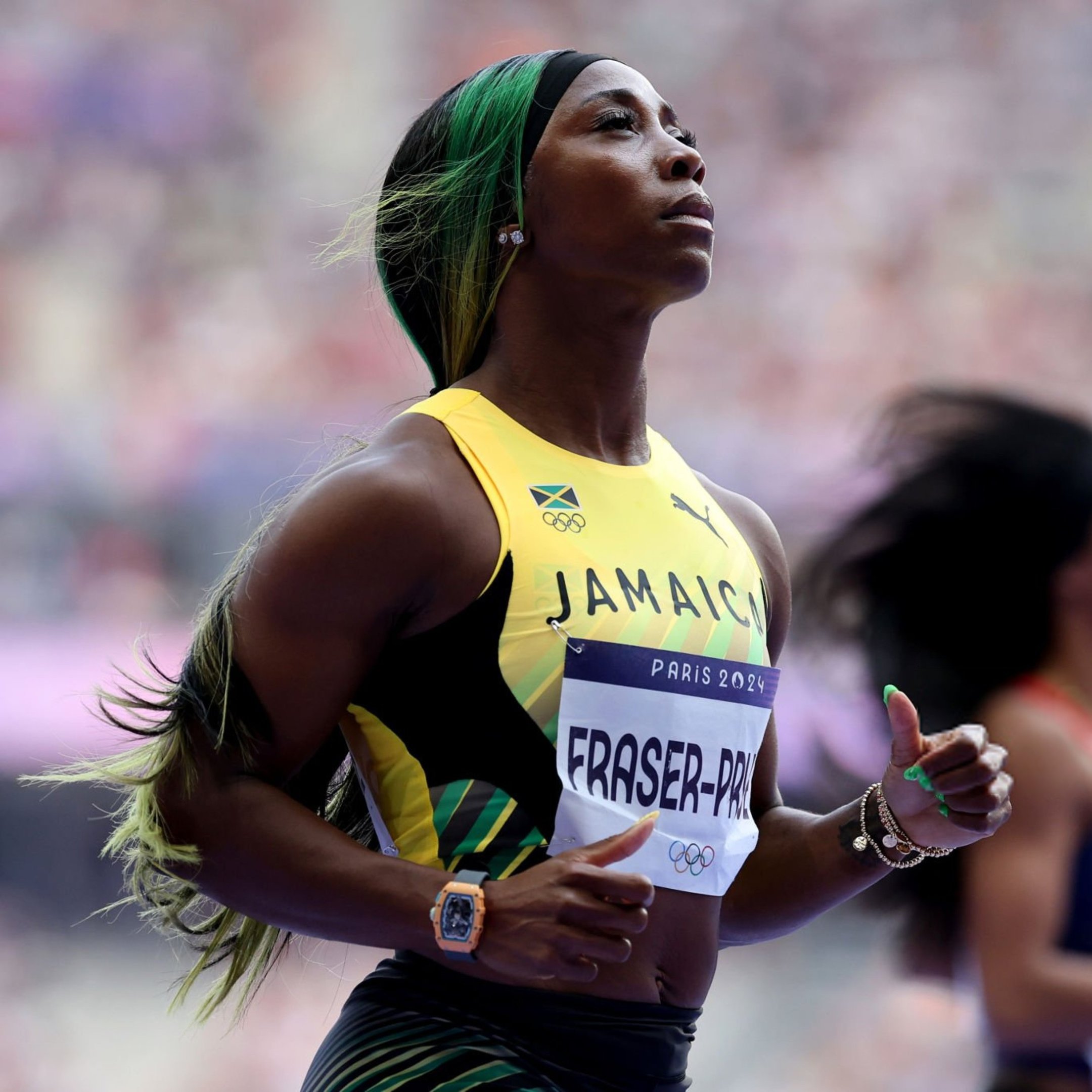 Atleta jamaicana vestindo uniforme esportivo com as cores verde, amarelo e preto. Usa acessórios como um relógio laranja e pulseiras. O cabelo está adornado com mechas verdes, seguindo a tendência de personalização de estilo em eventos esportivos.