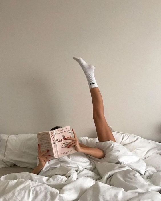 Pessoa deitada na cama sob lençóis brancos, lendo um livro com a perna estendida para cima, usando meias brancas de cano médio. Moda casual e confortável, estilo minimalista e descontraído. Tendência 