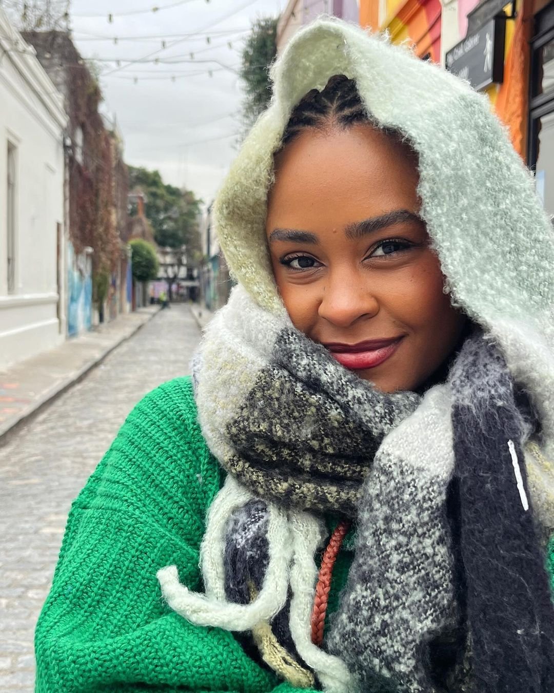 Pessoa com um look de inverno moderno e aconchegante. Usa um suéter verde de crochê combinado com um cachecol grande e macio em tons de preto, cinza e branco. Acessório de cabelo com tranças adiciona personalidade ao visual. hidratação da pele