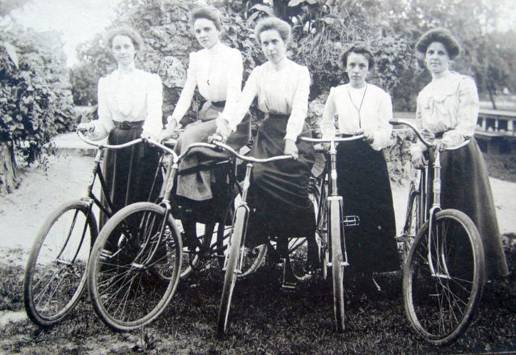 Imagem de cinco mulheres andando de bicicleta, vestidas com blusas de mangas compridas e saias longas, tendências de moda do início do século XX. Trajes formais e cabelos presos são destacados. Estilo vintage evidenciado pelo vestuário conservador.