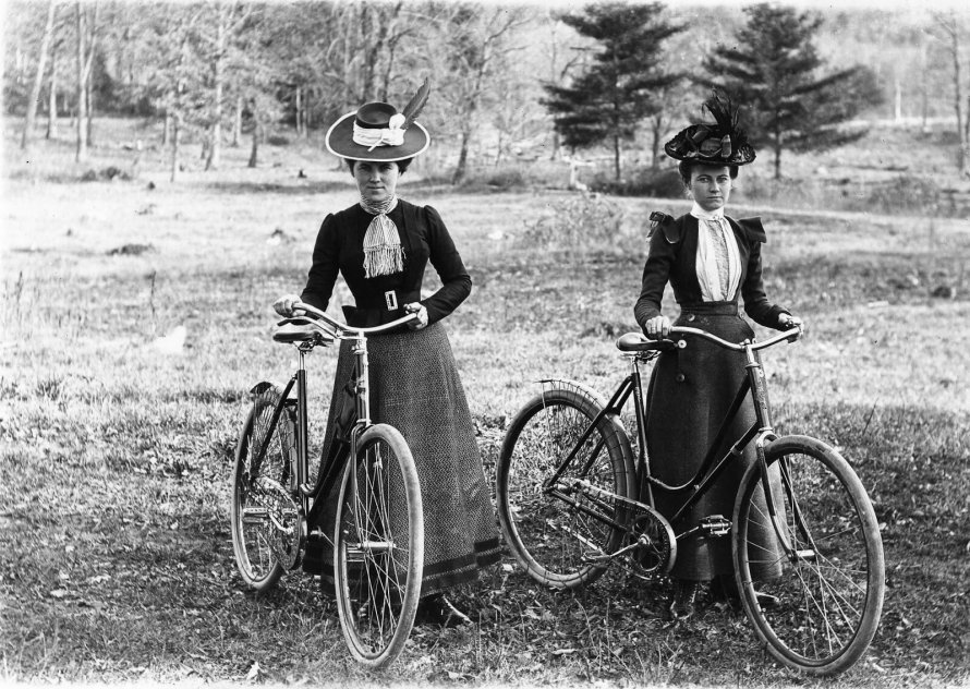 Duas mulheres com bicicletas vestem trajes da moda vitoriana, típicos do final do século XIX, com chapéus ornamentados, blusas de mangas puff e saias longas. O estilo reflete a elegância e o conservadorismo da época. Fundo de natureza.