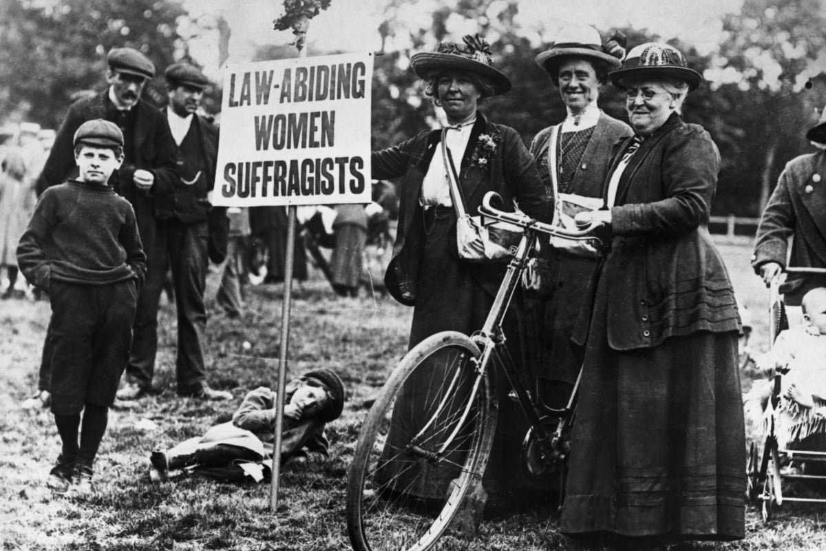 Mulheres sufragistas vestindo roupas típicas do início do século XX, como saias longas, blusas de gola alta, casacos com botões e chapéus adornados, seguram um cartaz durante uma manifestação. Homens e crianças ao fundo também apresentam trajes da época.