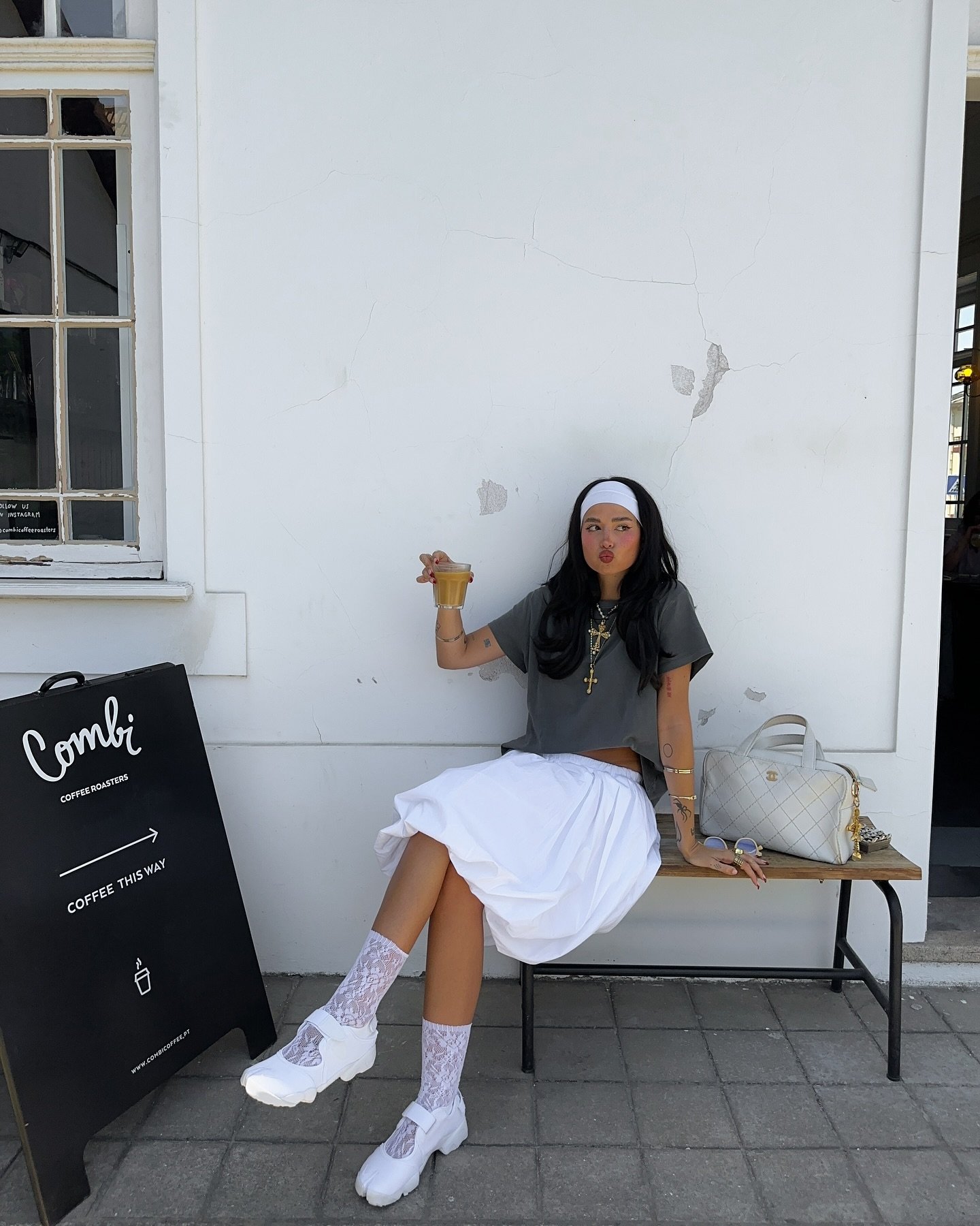 Jovem vestindo camiseta cinza cropped, saia branca ampla, meia rendada branca e tênis robusto também branco. Completa o look com faixinha branca, colares em camadas e bolsa de ombro clara.