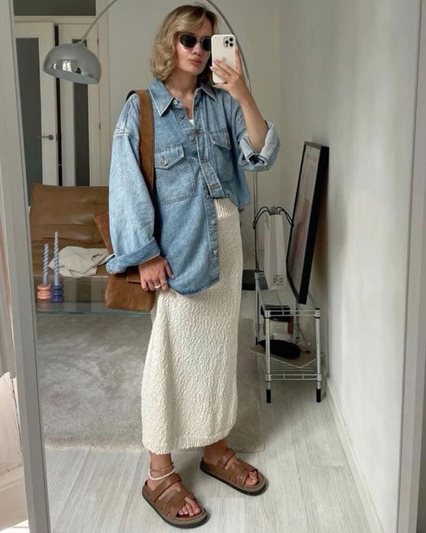 Look casual e moderno com camisa oversized jeans, saia midi texturizada branca, sandálias marrons estilo birken e bolsa de ombro marrom. Óculos de sol completam o visual.