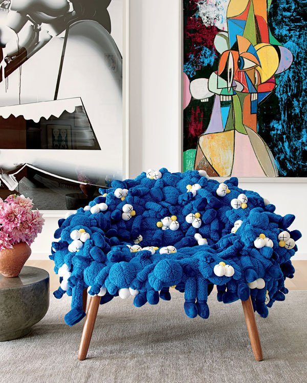 Cadeira moderna de pelúcia azul com detalhes brancos em estúdio de arte. Ao fundo, quadros contemporâneos com traços abstratos. A decoração mistura design arrojado e conforto, refletindo tendências de decoração ousadas e artísticas.