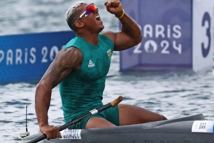 Atleta usando uniforme de remo verde, composto por regata e bermuda. Ele veste óculos de sol esportivos e exibe tatuagens nos braços. A combinação entre moda esportiva e acessórios funcionais reflete tendências de performance e estilo no esporte.