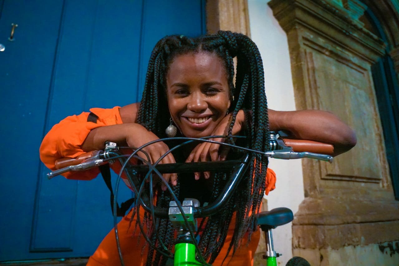 Mulher negra sorrindo, usando cabelos trançados longos e volumosos, vestido laranja vibrante, e brincos prateados grandes. Moda casual urbana, com foco em cores vivas e acessórios chamativos, posando sobre uma bicicleta verde em um ambiente noturno.