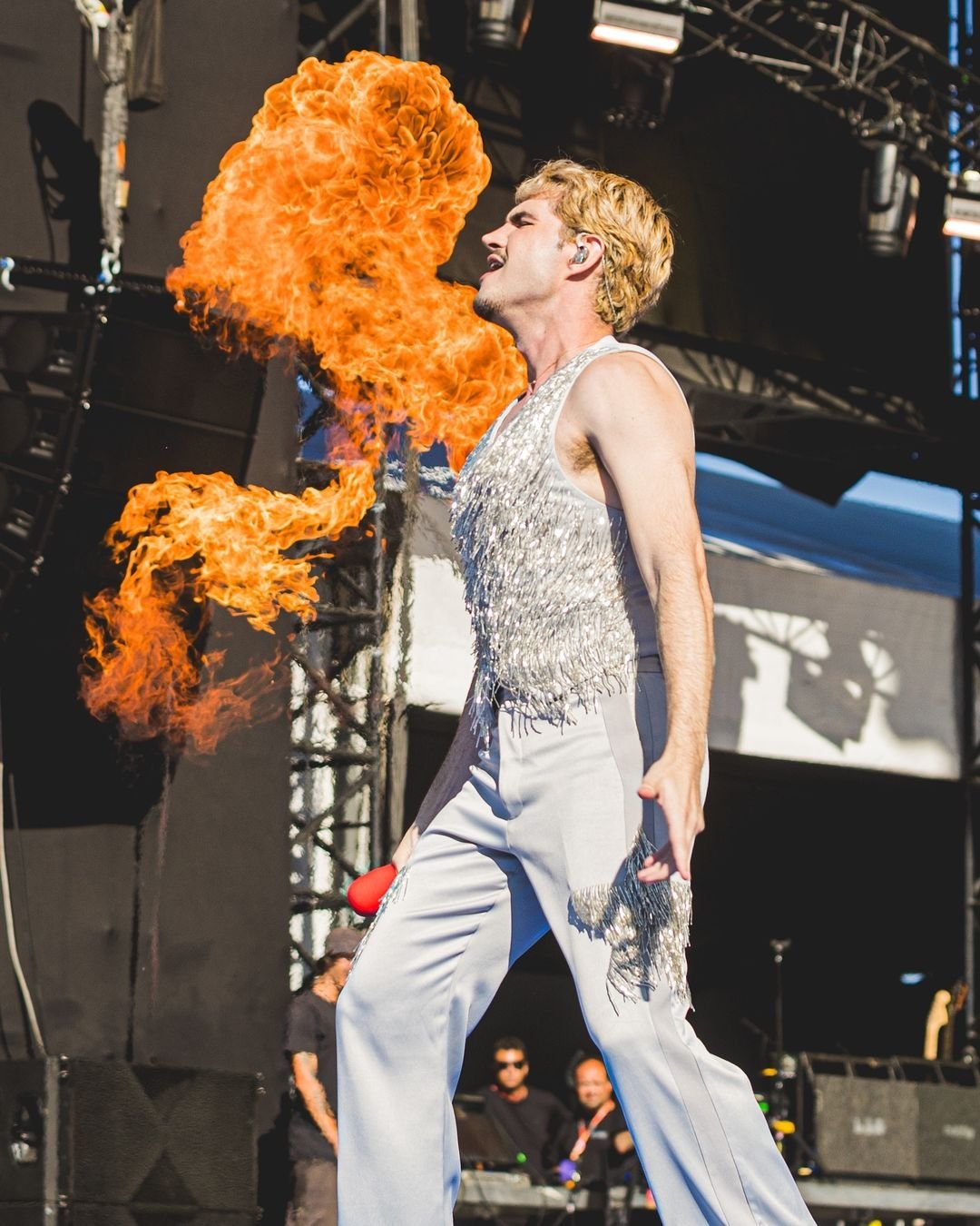 Artista em um show vestindo calça flare e colete prateado com franjas, inspirado na moda vintage dos anos 70. Tendência: Look retrô, com brilho e franjas, estilo glam rock. Efeito visual com fogo adiciona impacto à performance teatral.