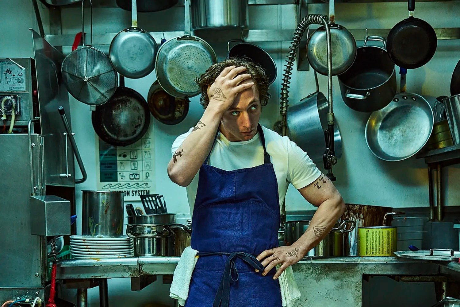 Chef usando camiseta branca básica e avental azul em uma cozinha profissional. Ele tem tatuagens nos braços e está cercado por panelas e equipamento de cozinha. Estilo descontraído e funcional, típico de cozinhas industriais.