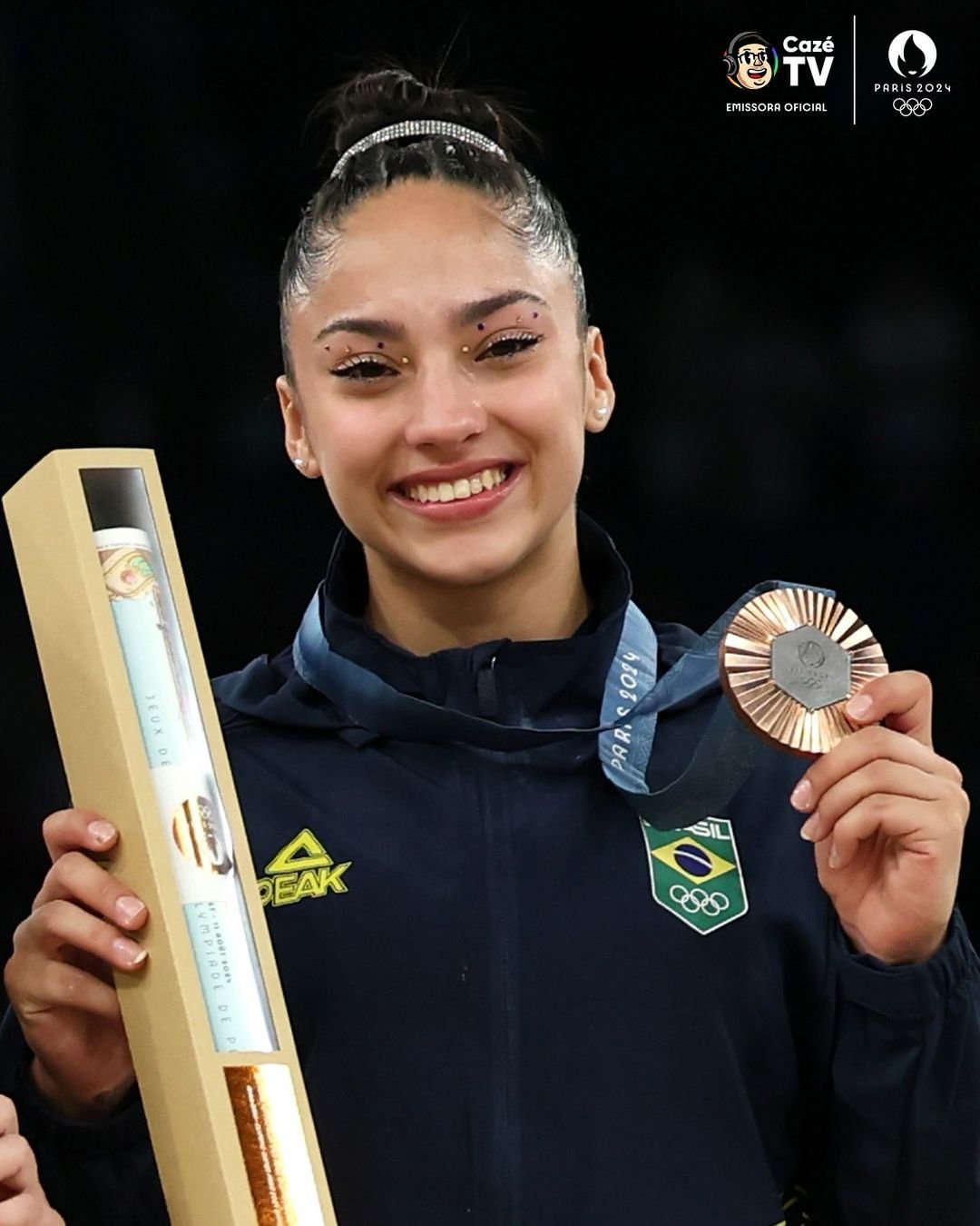 Atleta em traje esportivo, utilizando um agasalho azul marinho com logomarca do Brasil e PEAK. Maquiagem destacada com cristais ao redor dos olhos, cabelo preso em coque com tiara brilhante, transparecendo elegância e força na moda esportiva.